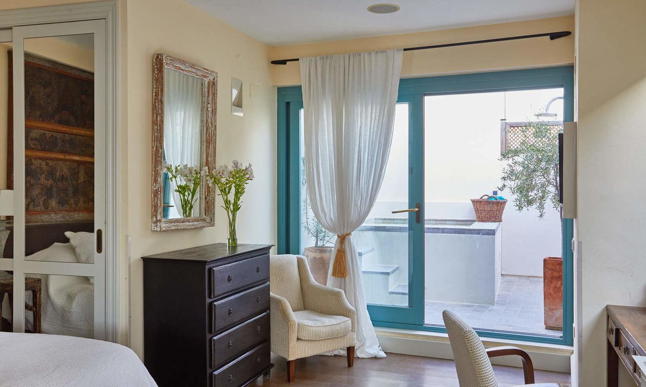 A bedroom with a dark chest of drawers, an upholstered armchair and a curtained glass door open onto a tiled terrace with a plunge pool and terracotta pots.