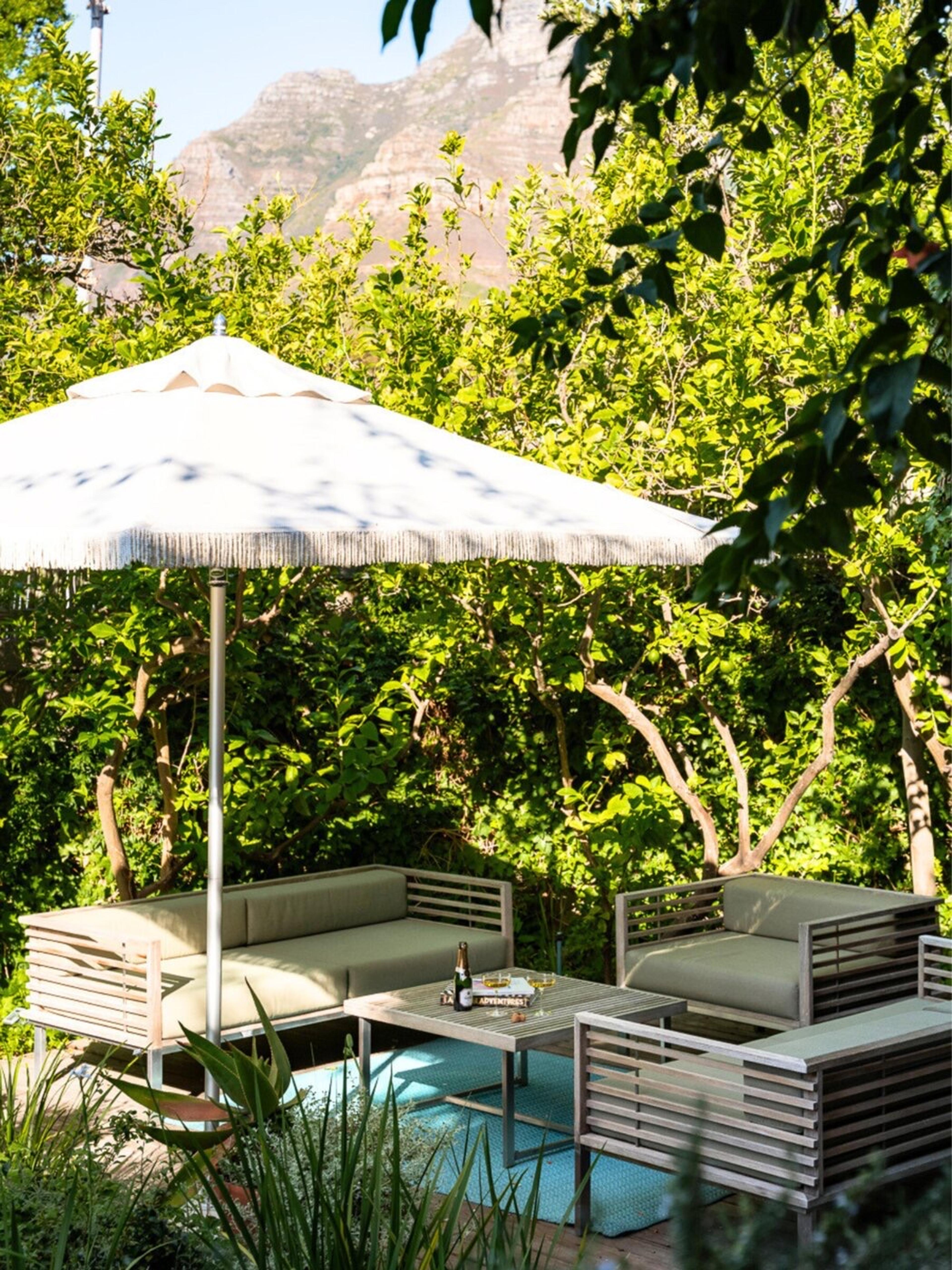 A shaded garden corner with a cream parasol over a grey slatted sofa and armchairs, a low table with a bottle and glasses, and fruit trees behind.