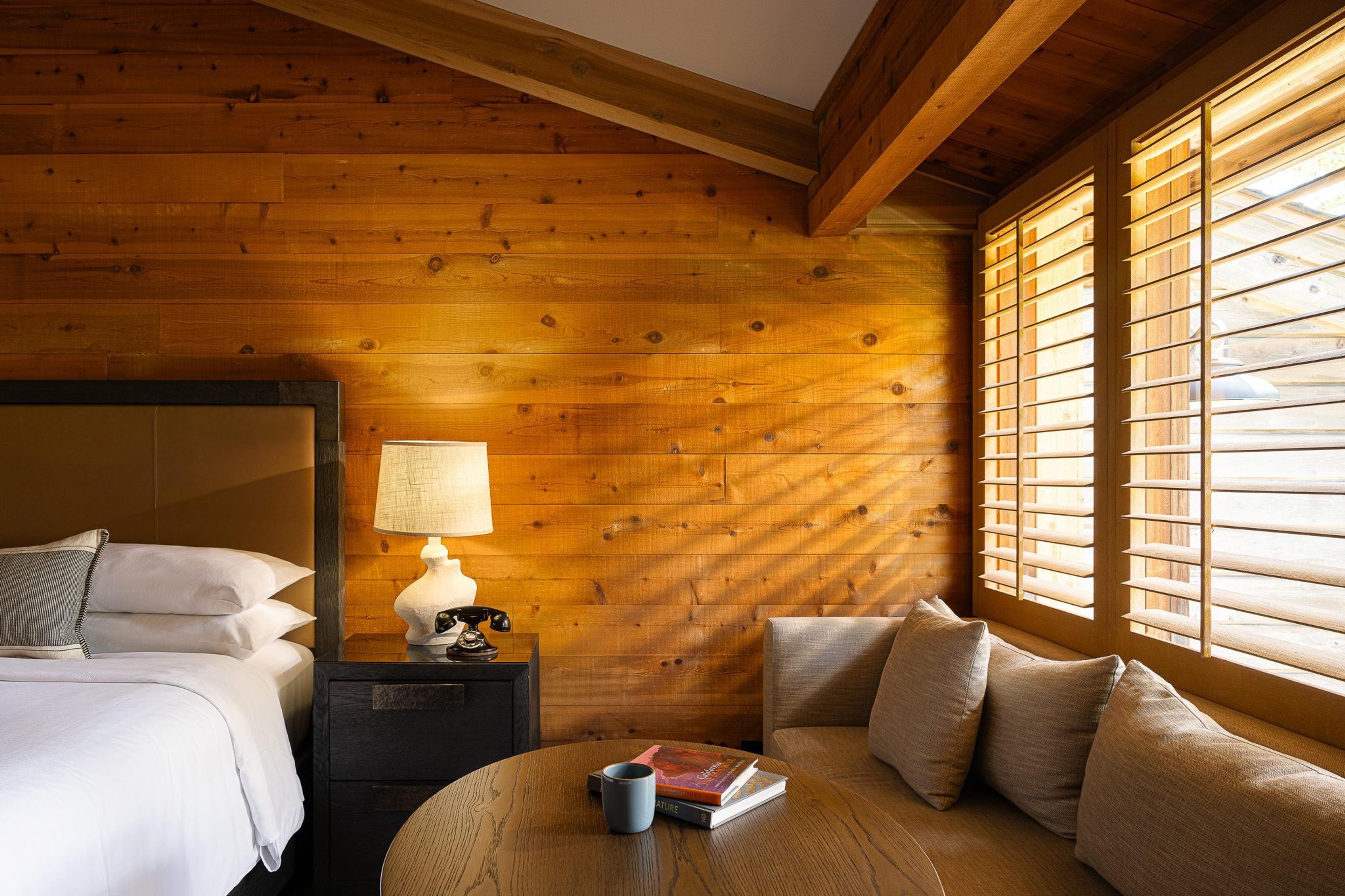 a wood-panelled bedroom with a bed, a bedside lamp, a small sofa and louvred shutters at the window