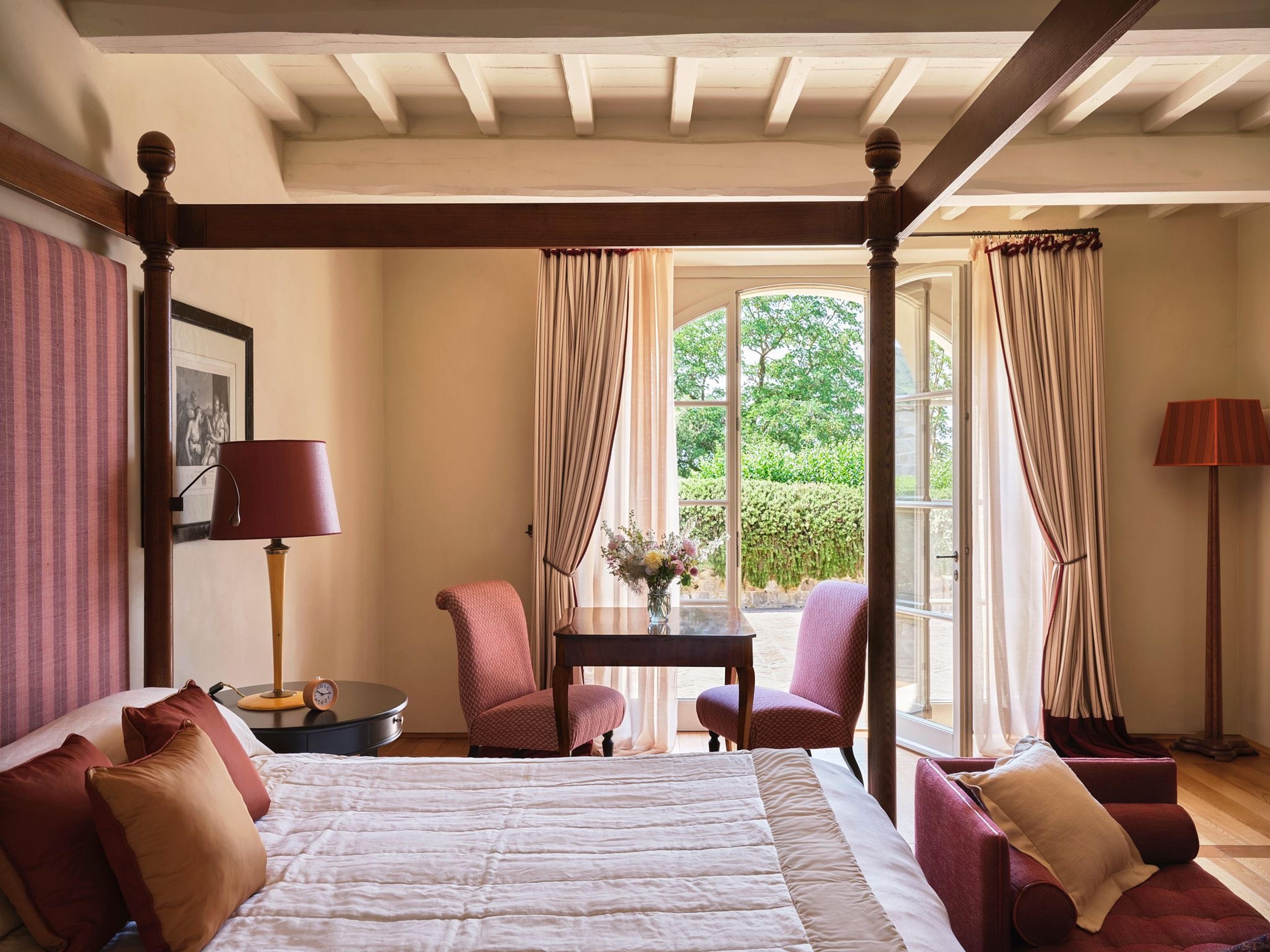 A four poster bed in a beamed bedroom, with armchairs by an open door leading to a garden.