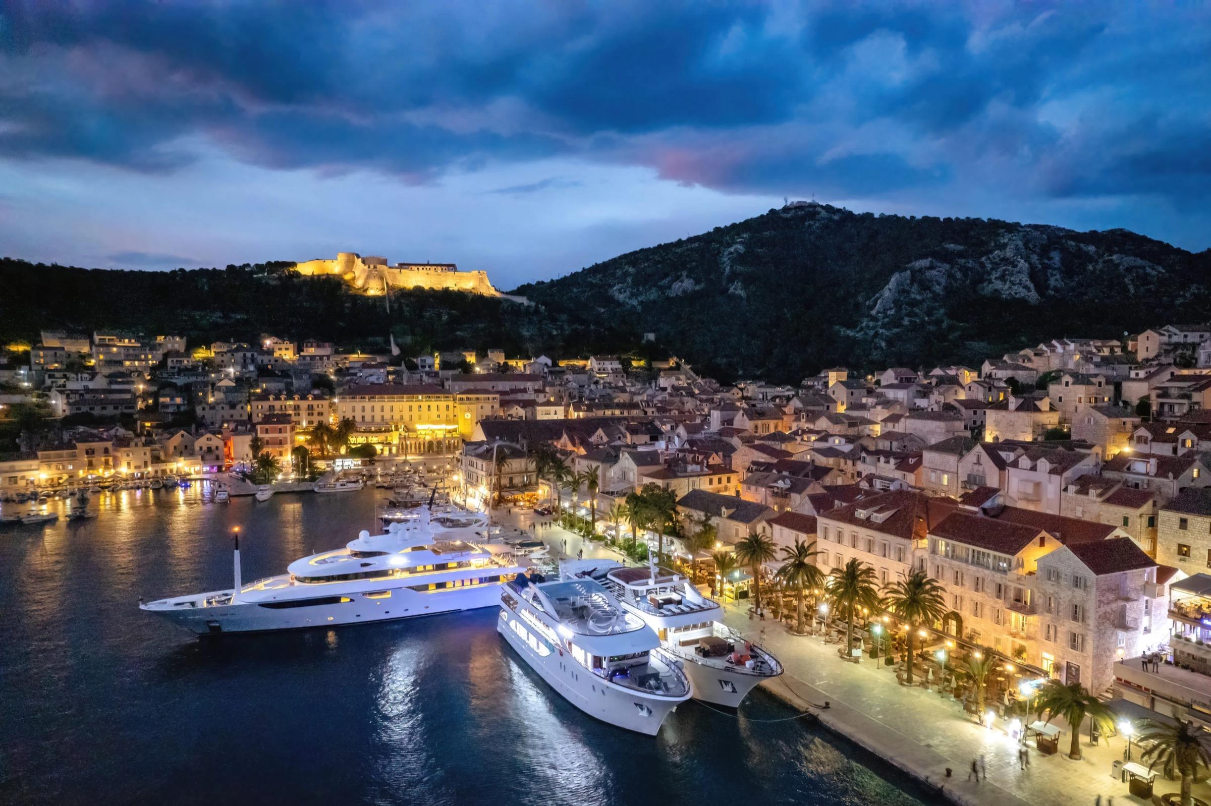 An aerial twilight view of a harbour town with a hilltop fortress, two superyachts moored along a promenade, and lit streets.