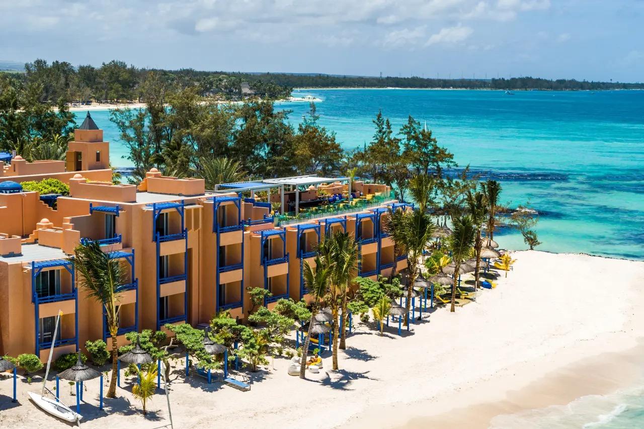 An aerial view of the hotel's terracotta wings along a white sand beach, with a sailboat on the shore and a turquoise lagoon beyond.