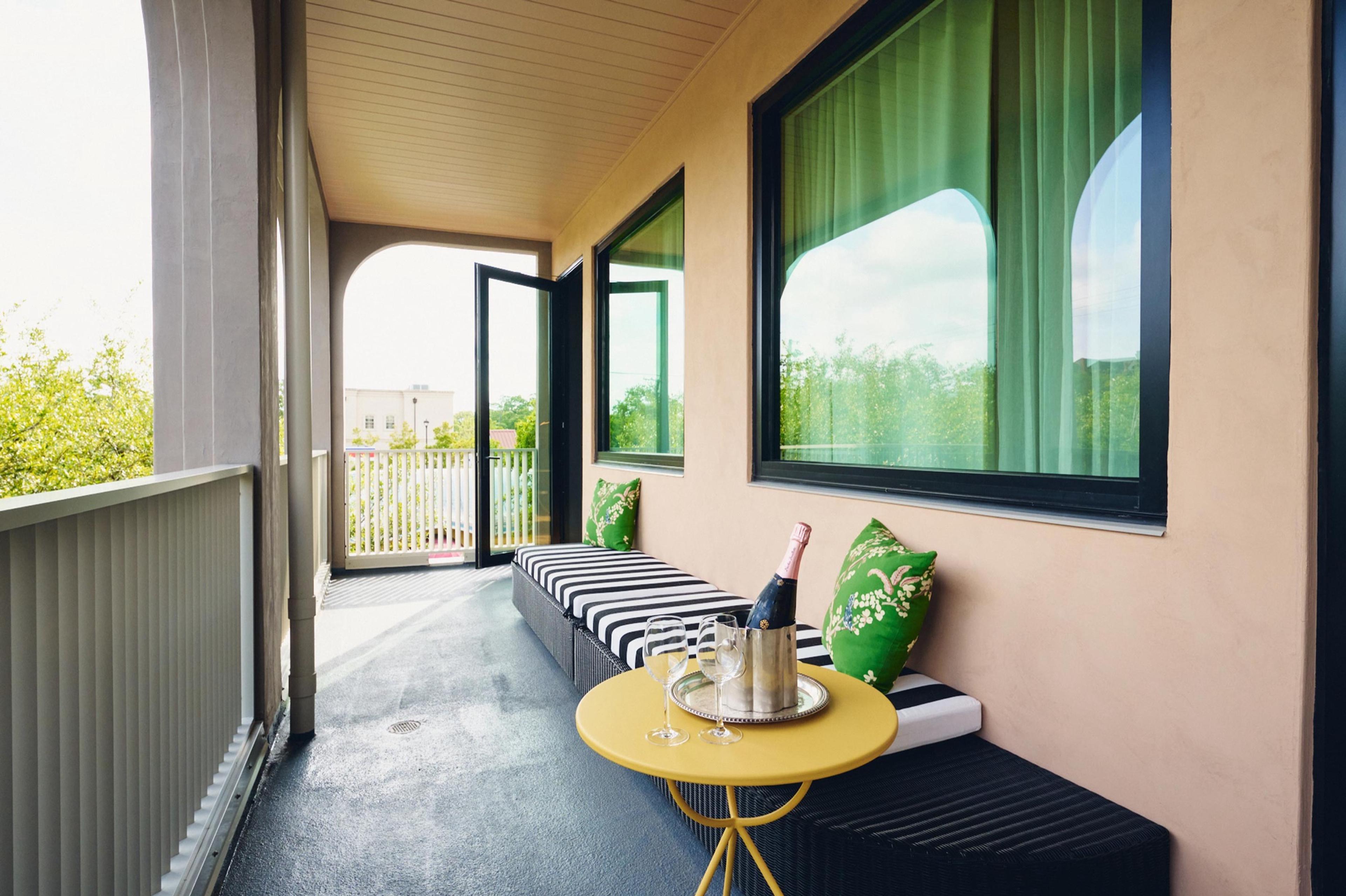 A balcony with a striped bench, green cushions, a yellow table holding a champagne bottle in an ice bucket and two glasses, and trees beyond the railing.
