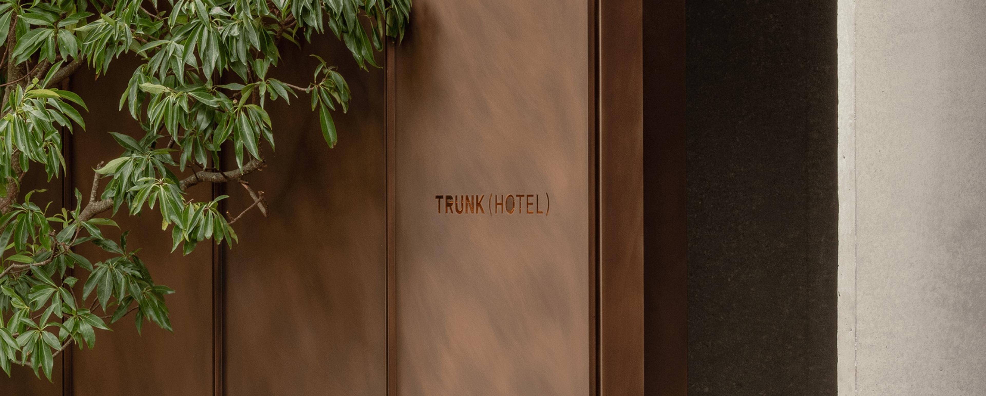 a weathered copper entrance door stamped Trunk (Hotel), with a leafy tree branch reaching across it