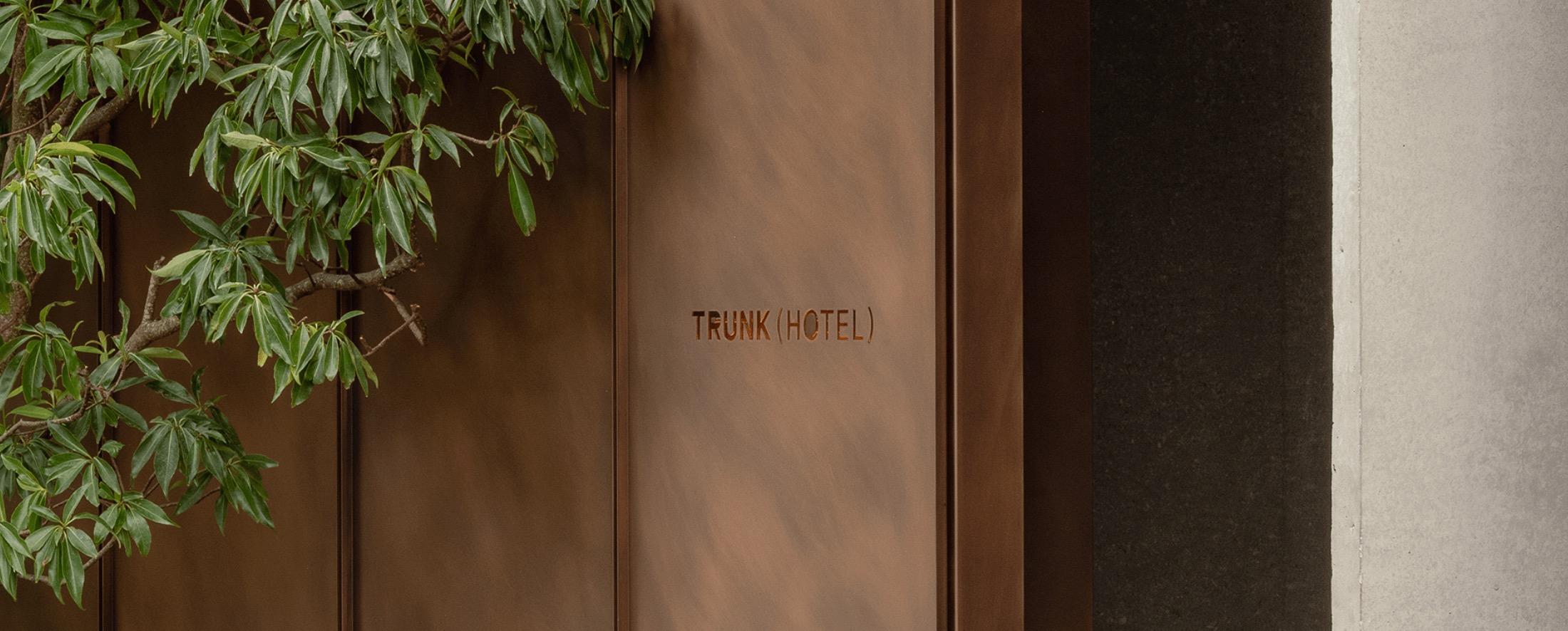 a weathered copper entrance door stamped Trunk (Hotel), with a leafy tree branch reaching across it