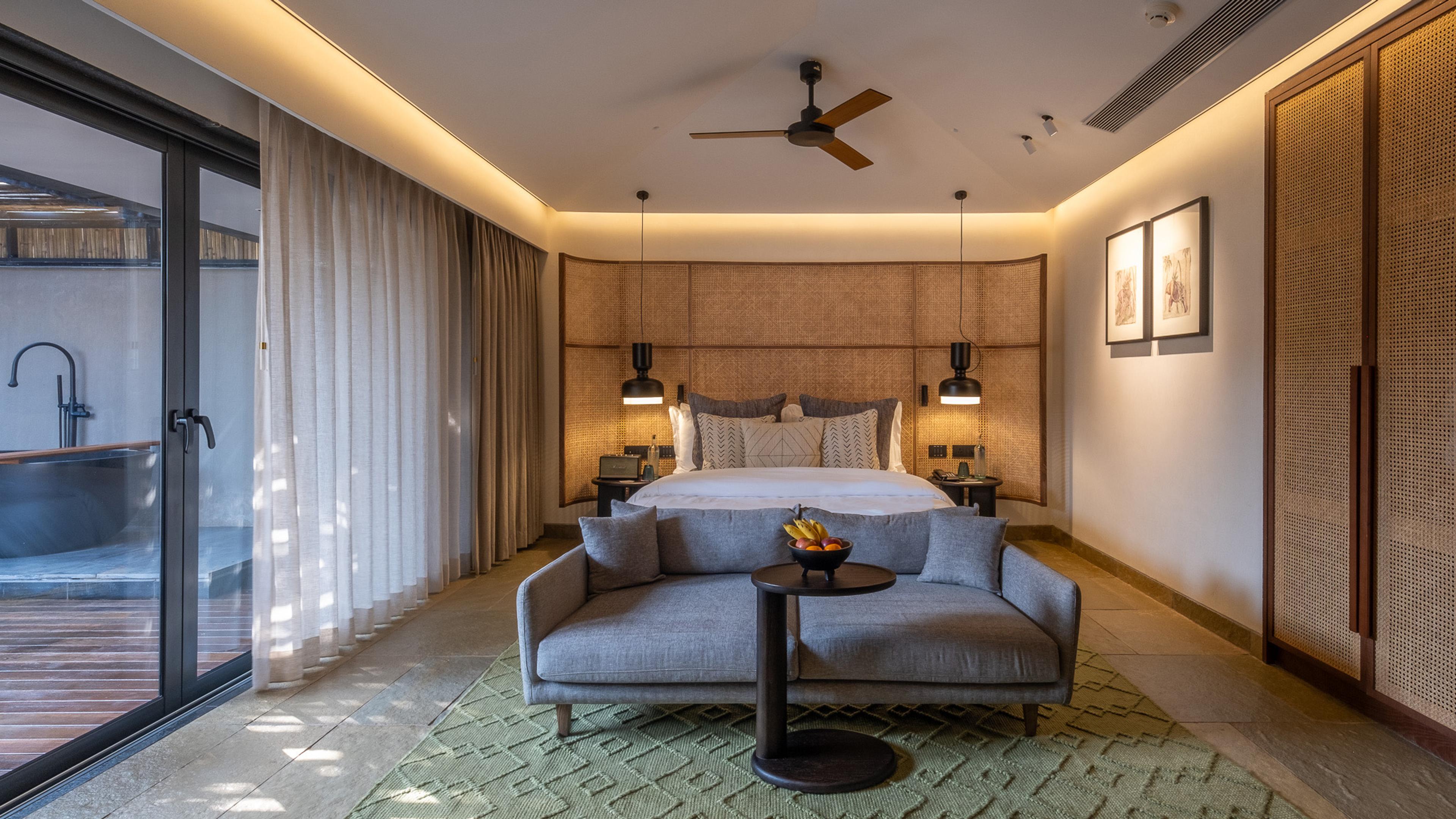 A bedroom with a grey upholstered headboard and framed prints, its glass doors open onto a deck with a freestanding bathtub.