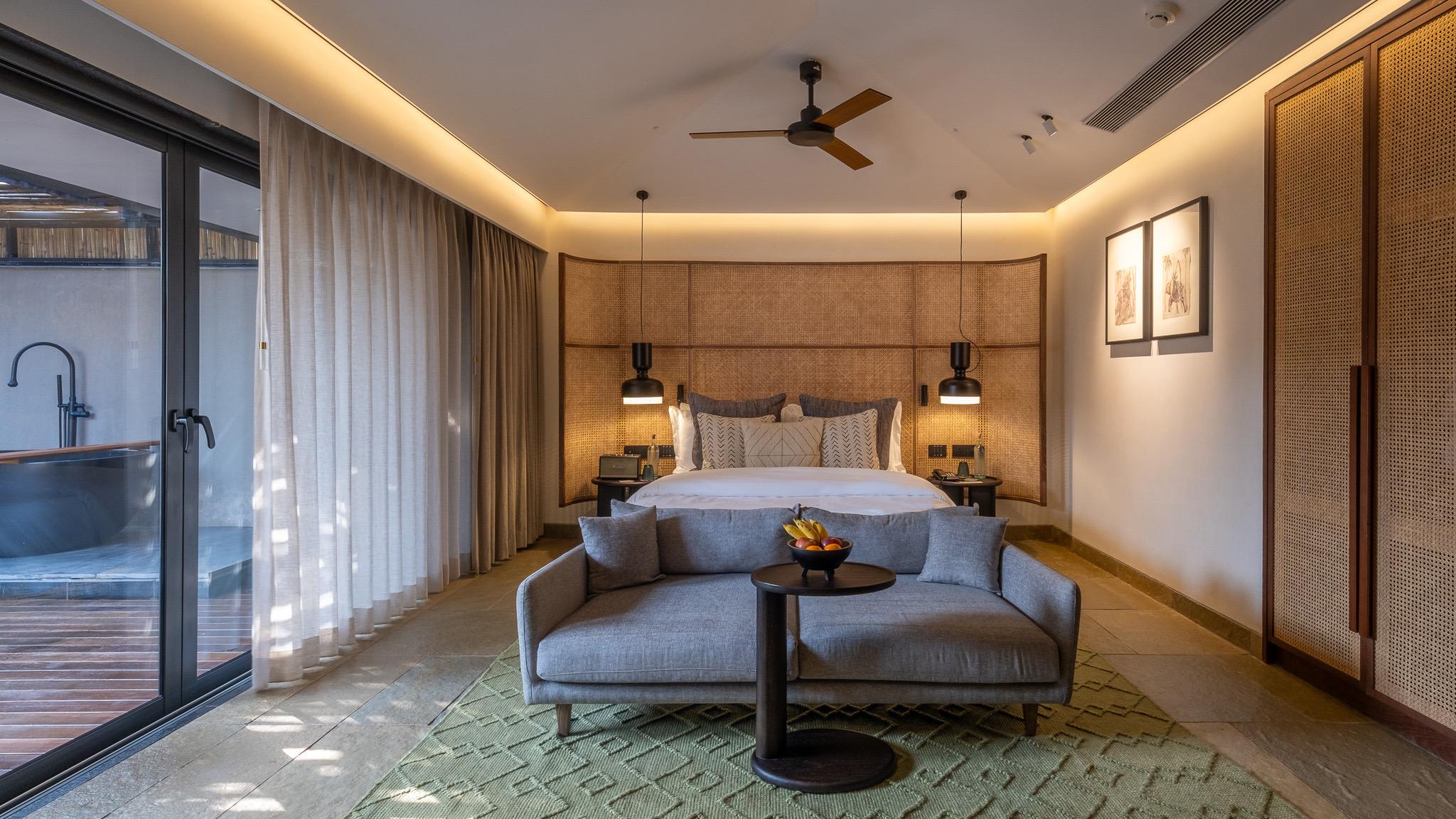 A bedroom with a grey upholstered headboard and framed prints, its glass doors open onto a deck with a freestanding bathtub.