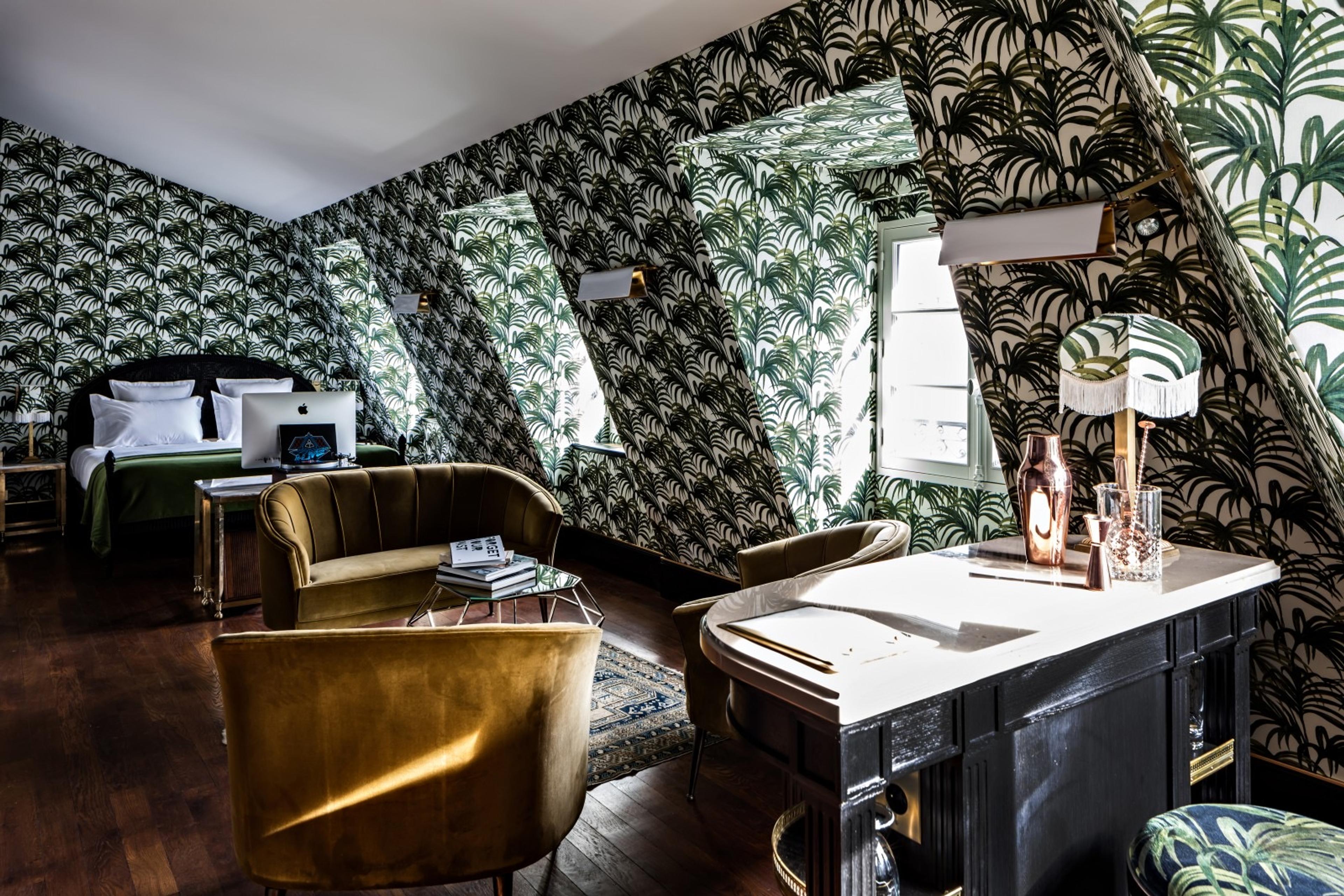 A sitting area with mustard velvet armchairs and a black desk, in a room with sloped ceilings covered in green palm-leaf wallpaper, a green bed visible in the background.