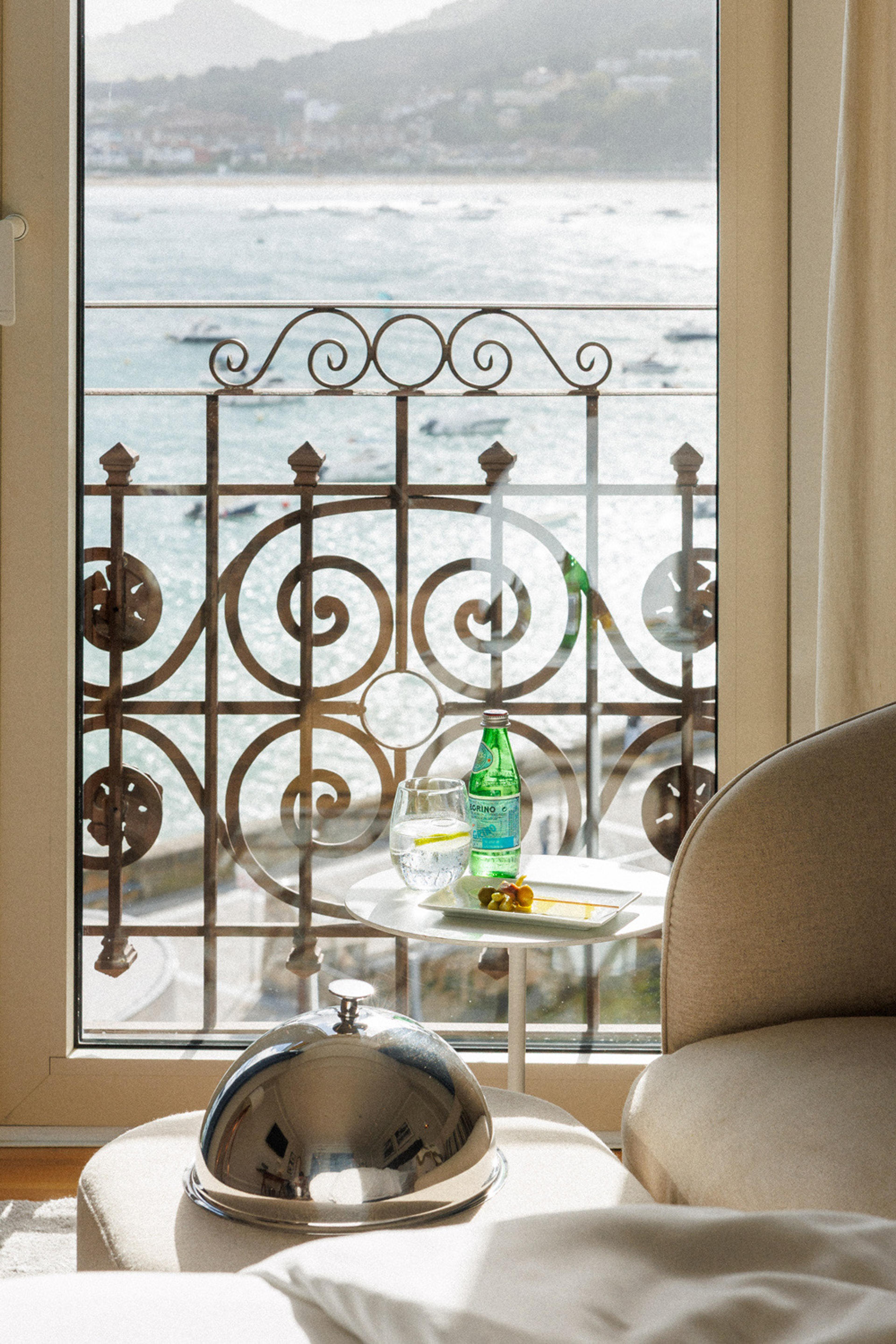 A table with a water bottle, a glass and a silver dish cover beside a wrought iron balcony rail looking over a harbour.