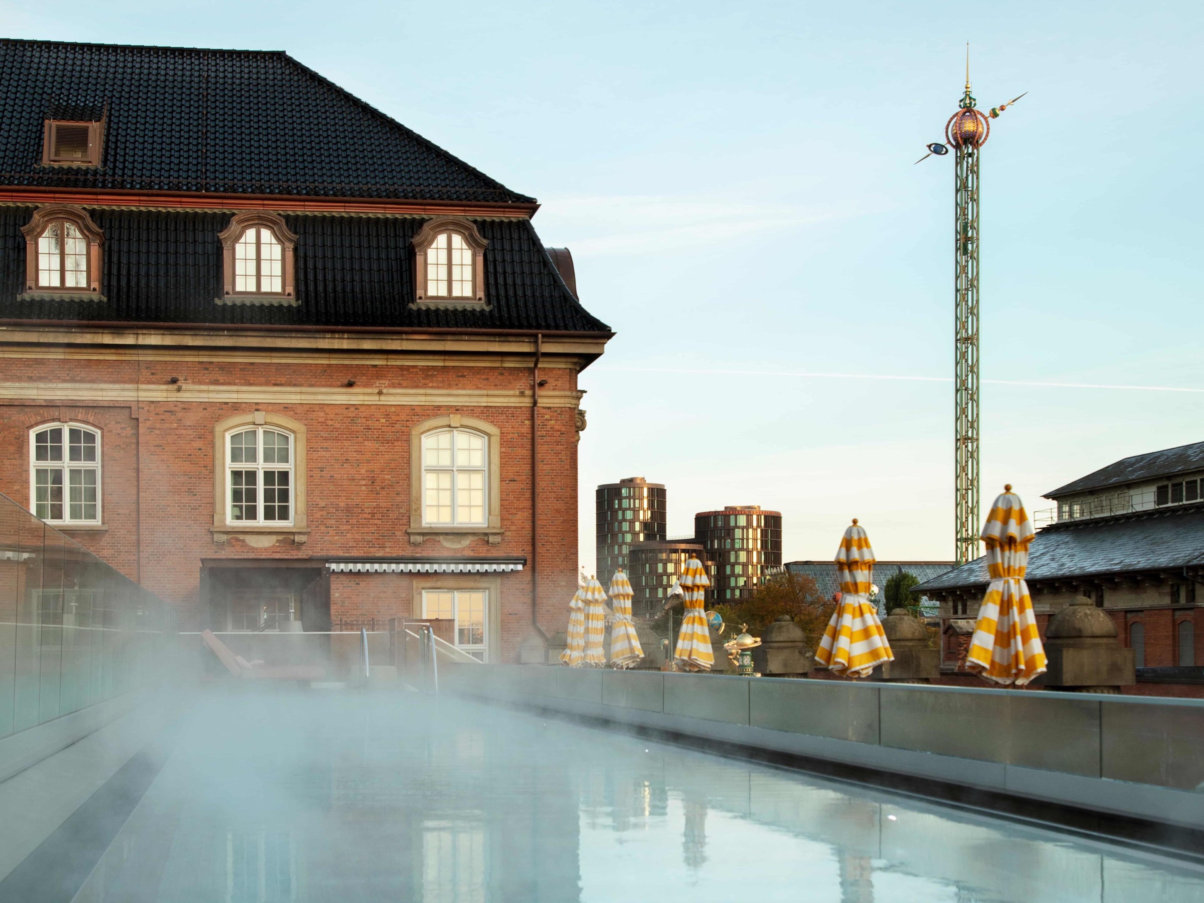 Steam rising off the rooftop pool at Villa Copenhagen, the red-brick old post office beside it and a Tivoli ride's tower beyond the striped parasols