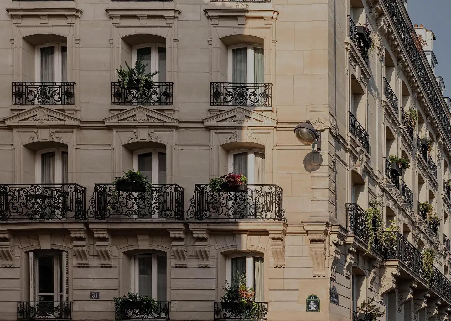 The stone facade of a Haussmannian corner building, with wrought iron balconies, window boxes and climbing plants, photographed from the street in daylight.