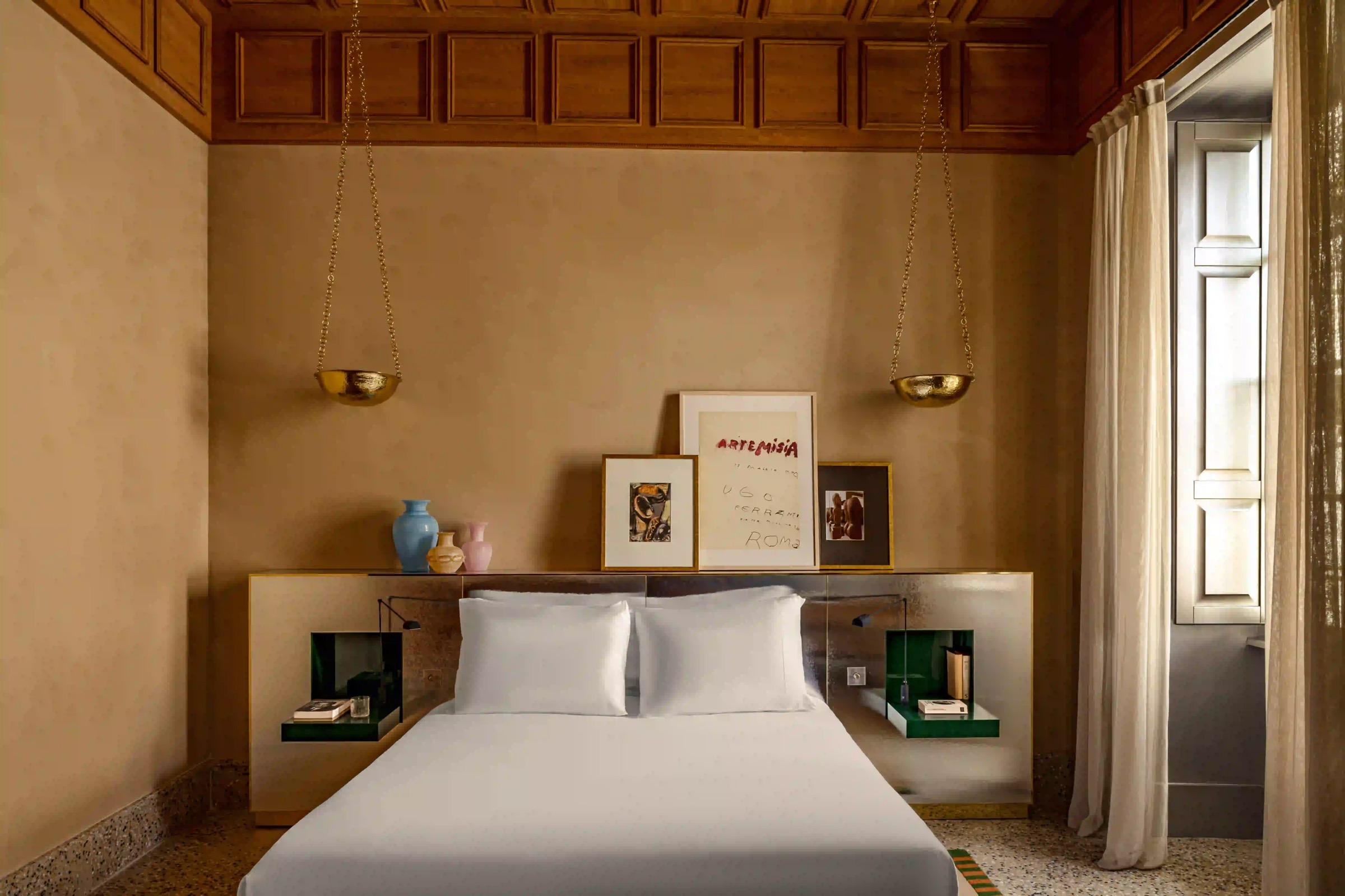A symmetrical bedroom with a white-made bed under a wood-panelled ceiling, framed artwork and small vases on the headboard shelf, and two brass bowl pendant lights hanging on chains.