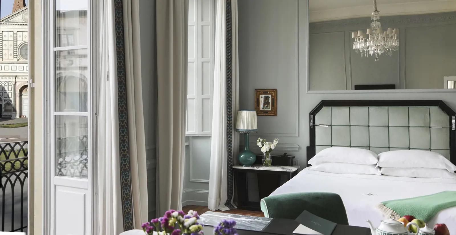 A bedroom with a chandelier reflected in a tall mirror, a bed with a quilted pale green headboard, and open glass doors leading to a balcony overlooking a church facade.