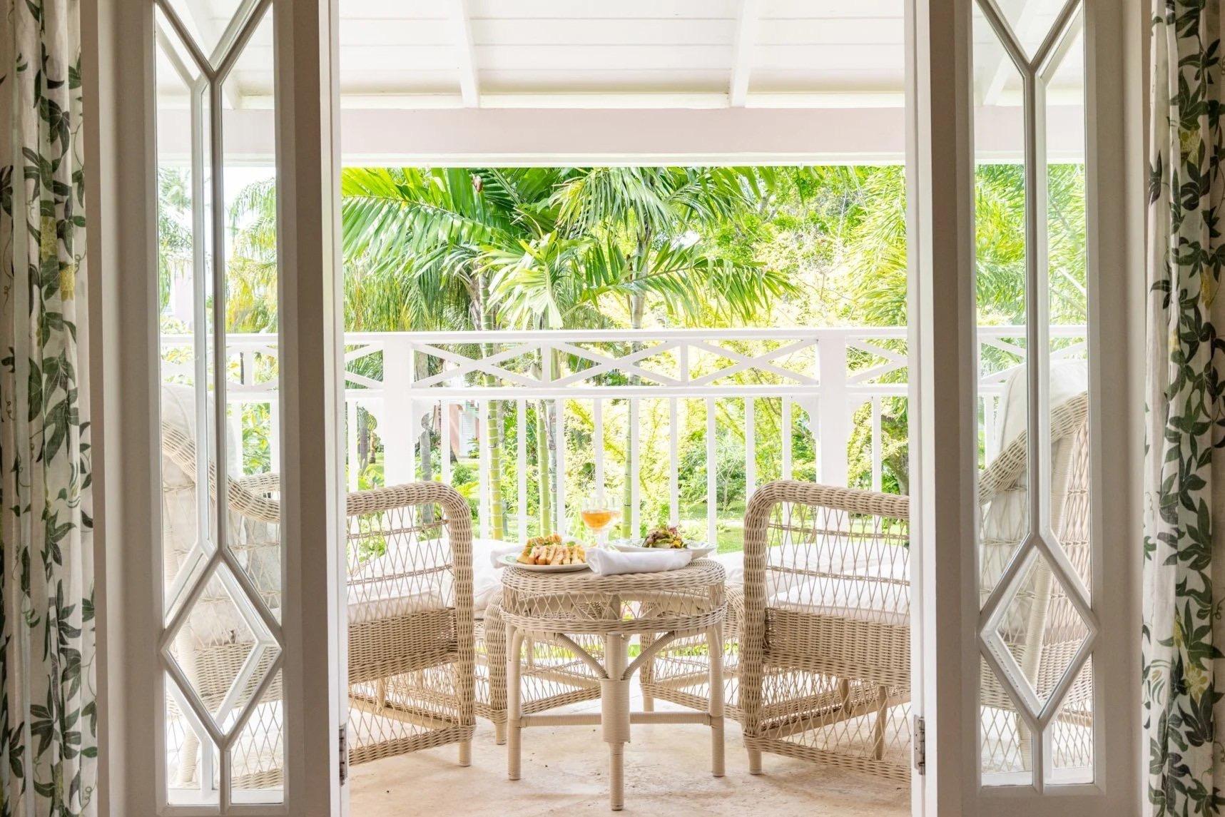 wicker chairs around a small table set with canapes and a cocktail, seen through open doors onto a balcony with palm trees beyond