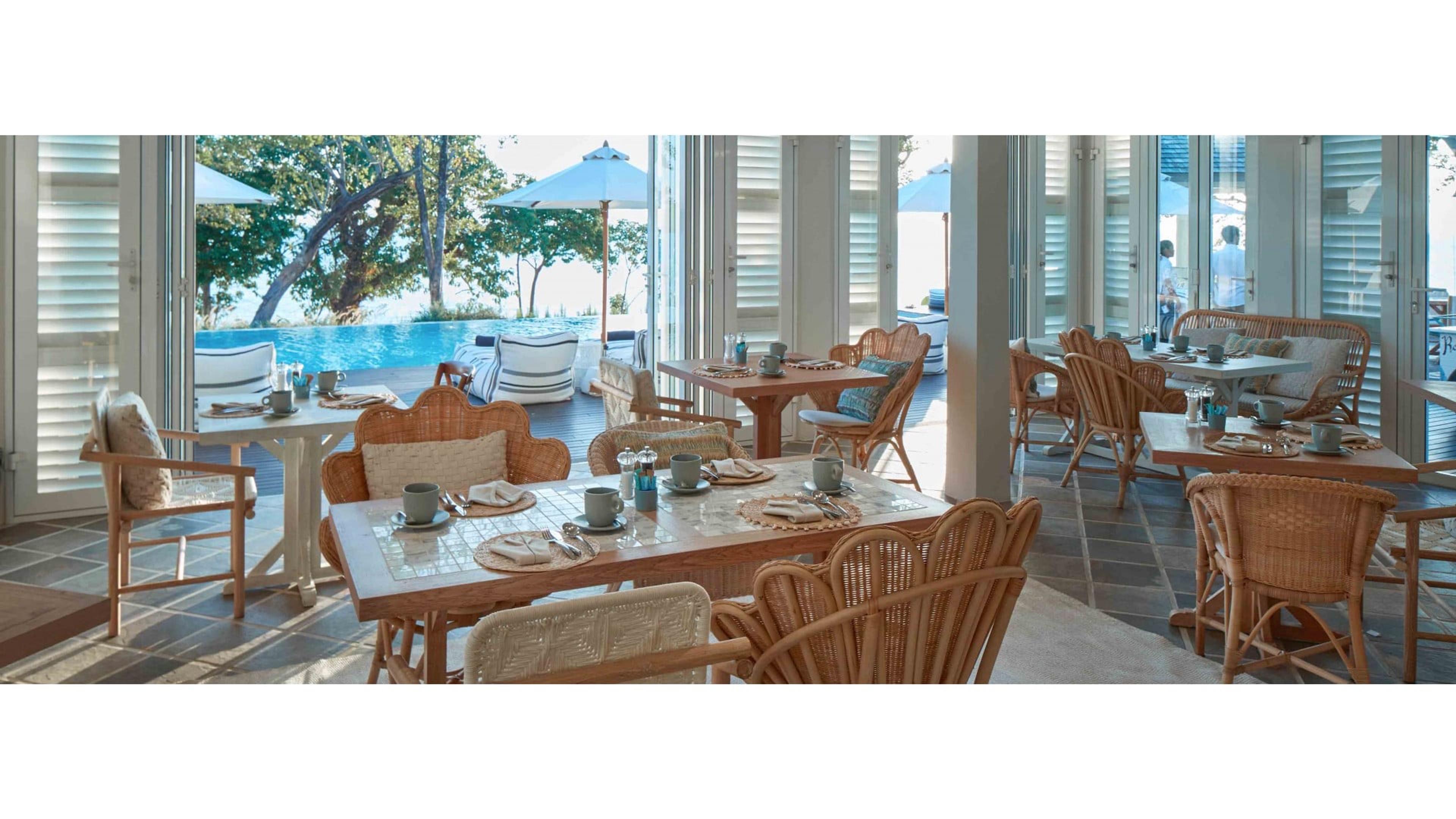 A dining room with rattan chairs and tables laid for breakfast, French doors open to a pool and the sea beyond, two people visible in the distance.