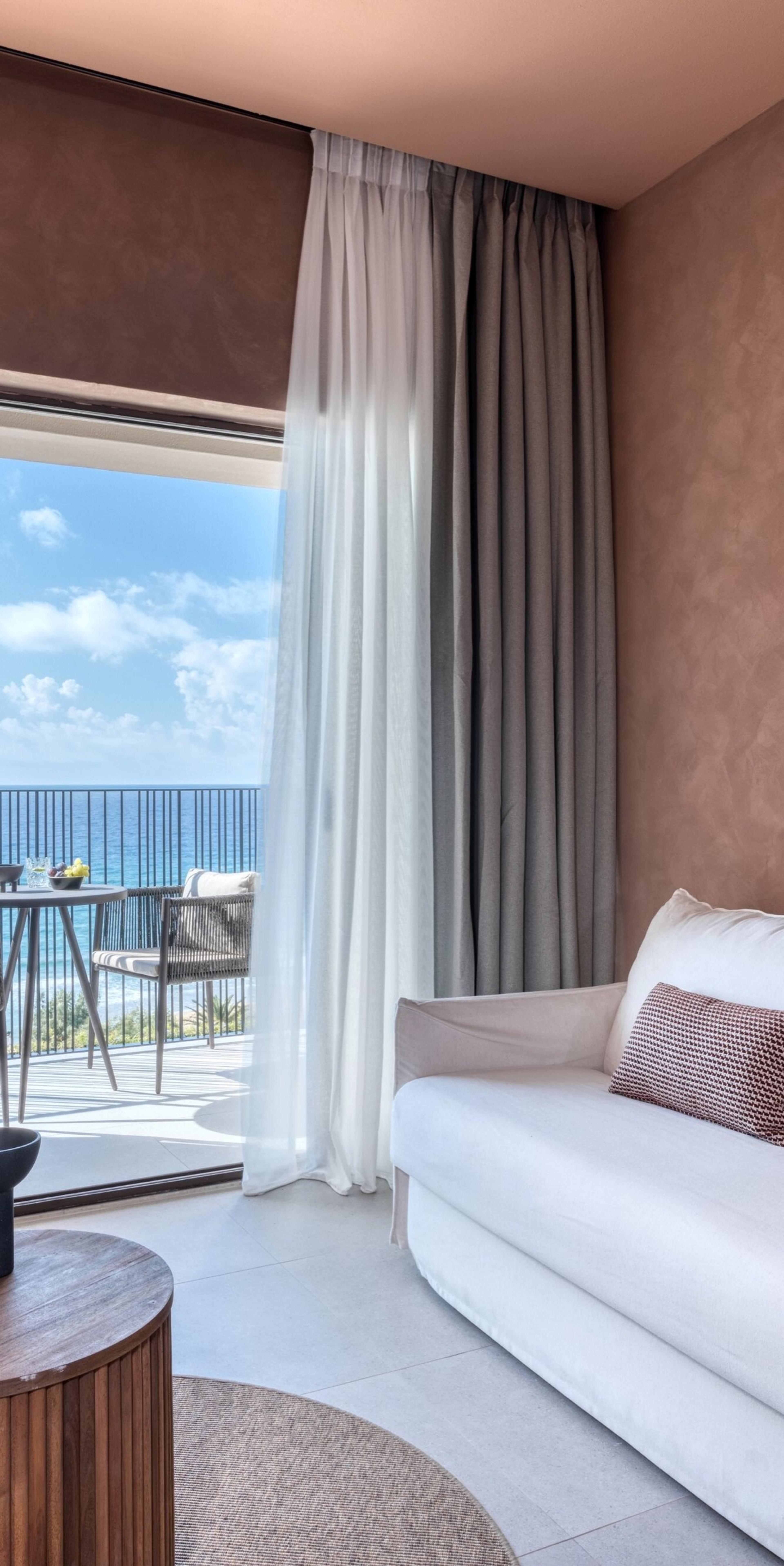 A white sofa with a patterned cushion sits beside sheer curtains, looking through an open balcony door to a table, chairs, and the sea.