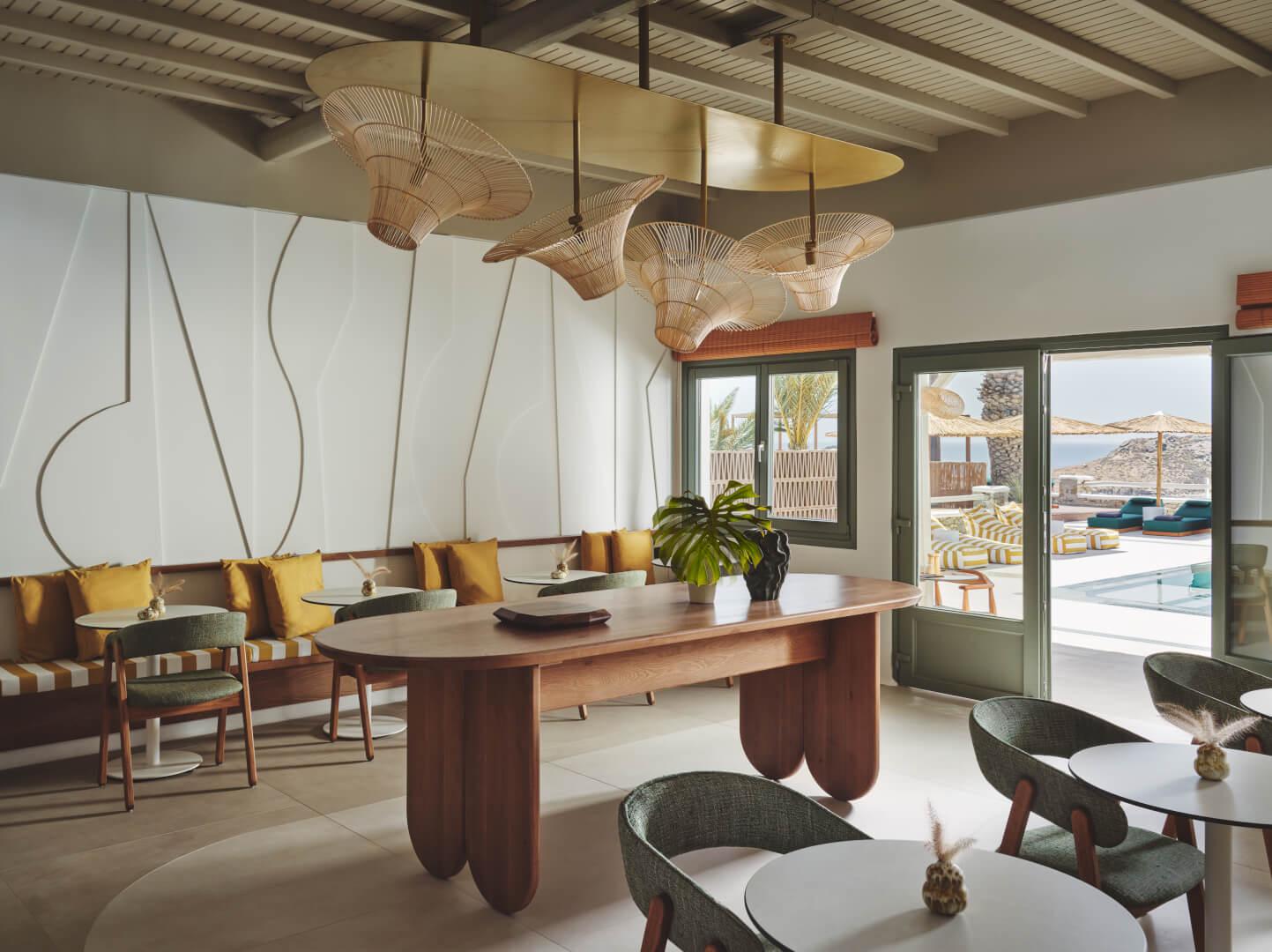 An empty dining lounge with a long wooden table, green chairs, striped banquette seating, and woven pendant lights, with a pool visible through open doors.
