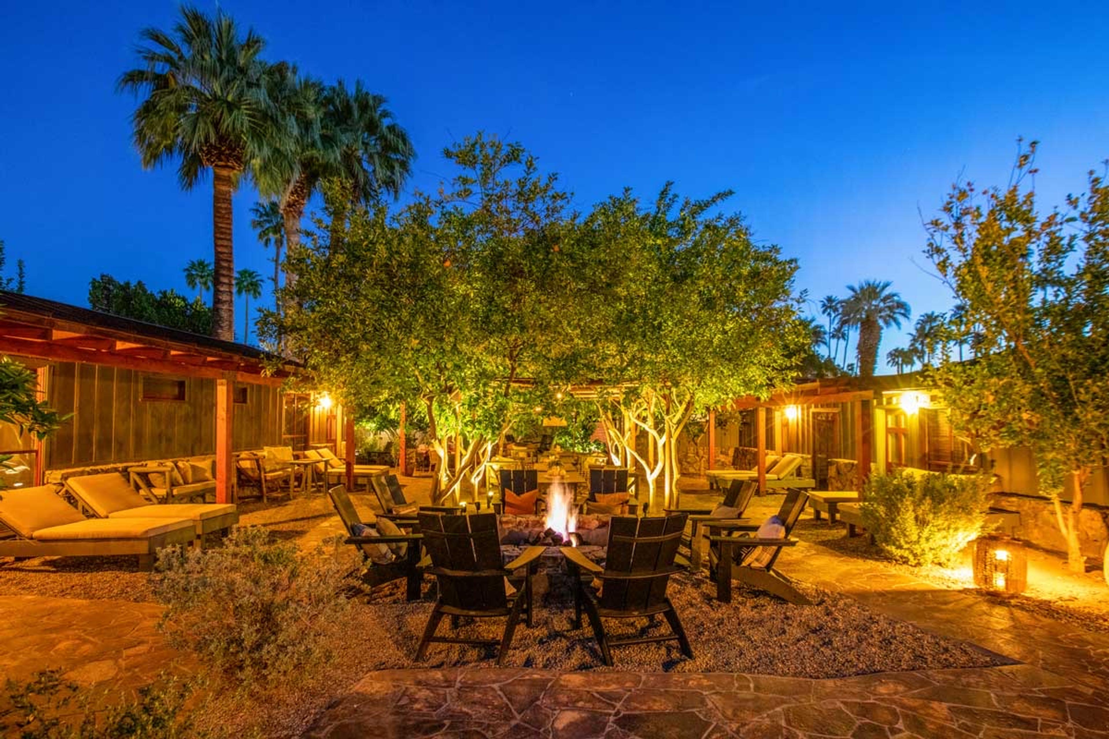 A courtyard at dusk with a lit fire pit at its centre, ringed by wooden Adirondack chairs, under trees lit from below, with low buildings and loungers around the edges.