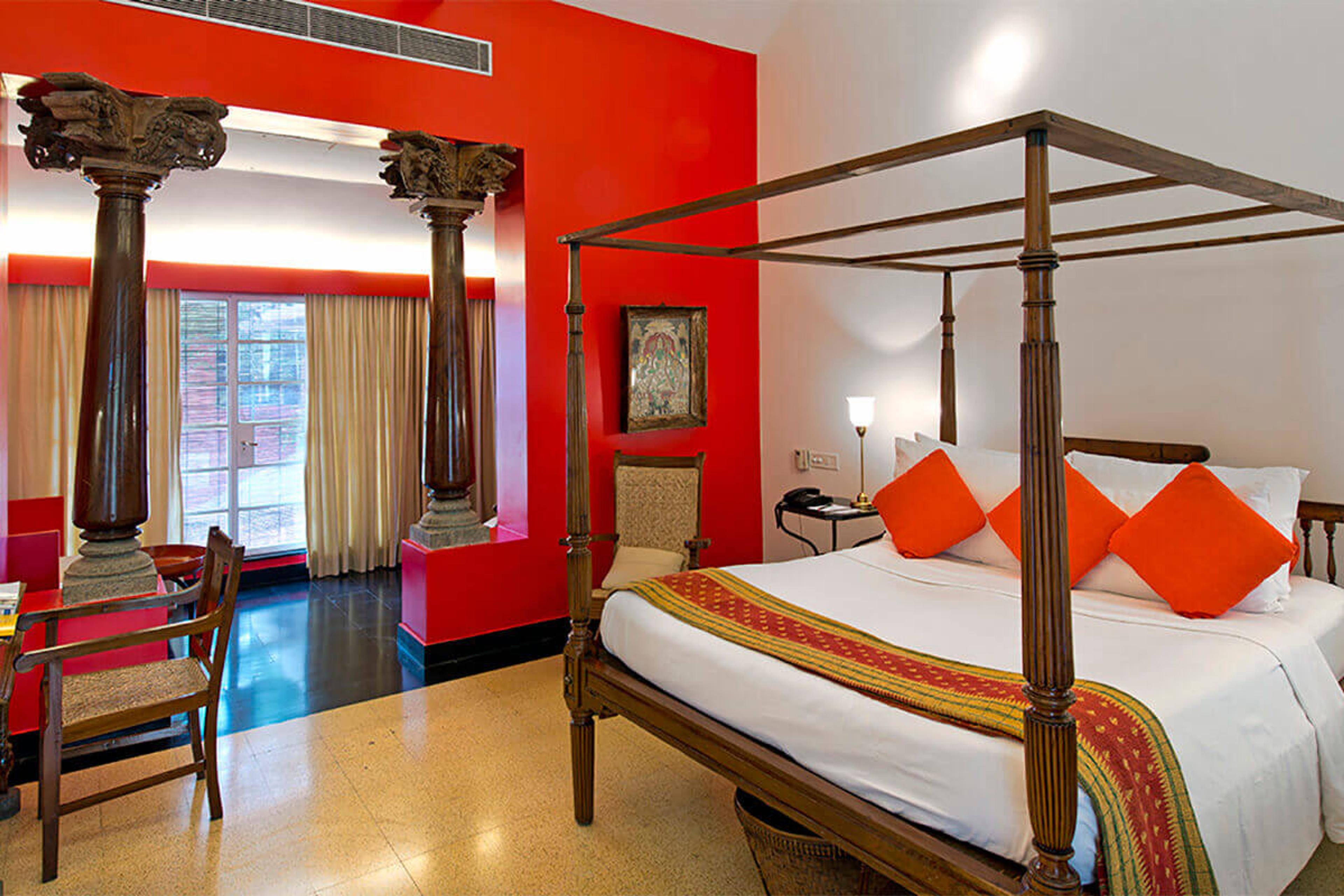 A red-walled bedroom with a dark wood four-poster bed dressed in white linen and orange cushions, seen through a pair of carved pillars.
