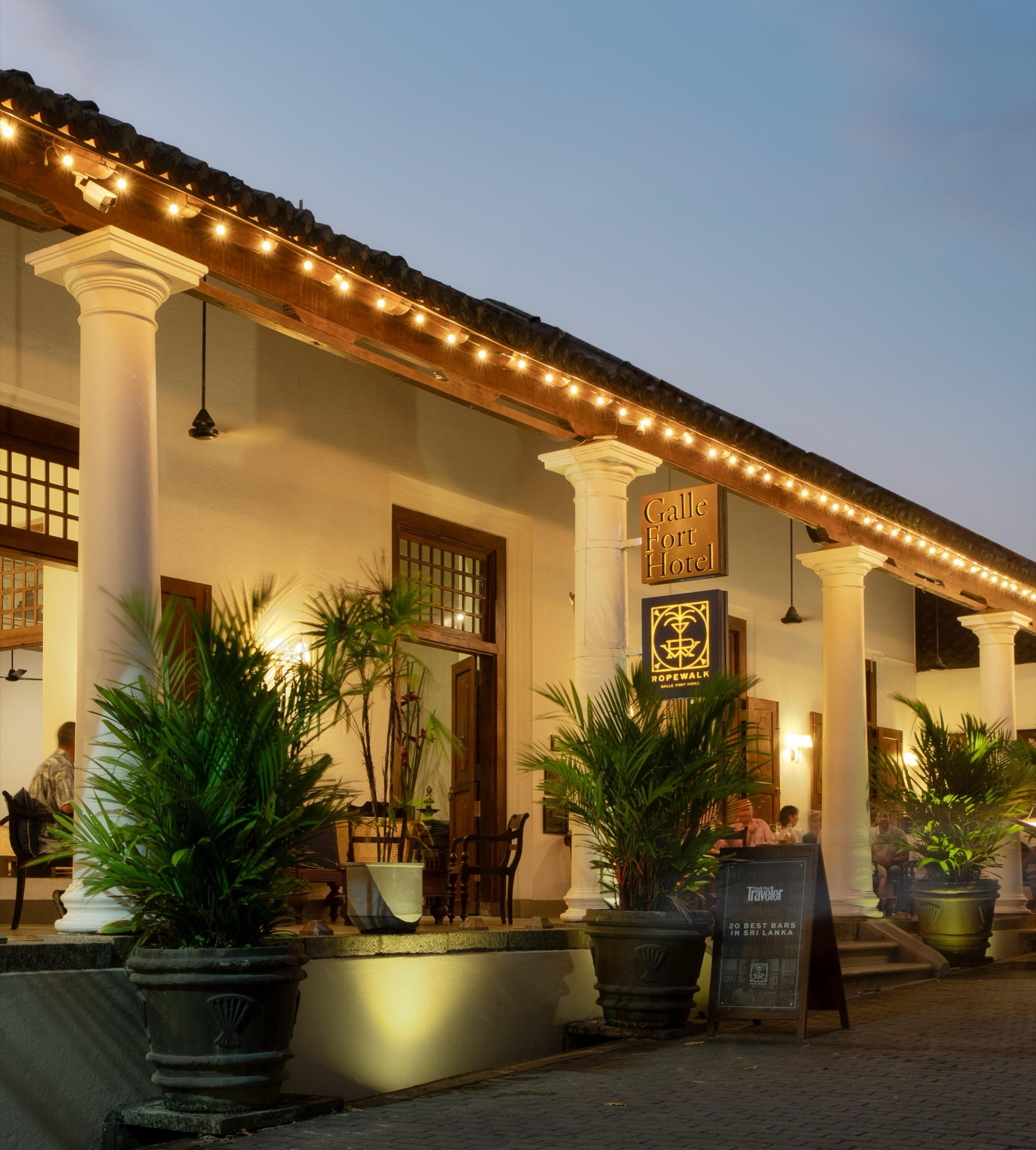 The hotel's colonnaded front at dusk, strung with small lights, with potted palms on the terrace and guests seated at tables in the background.