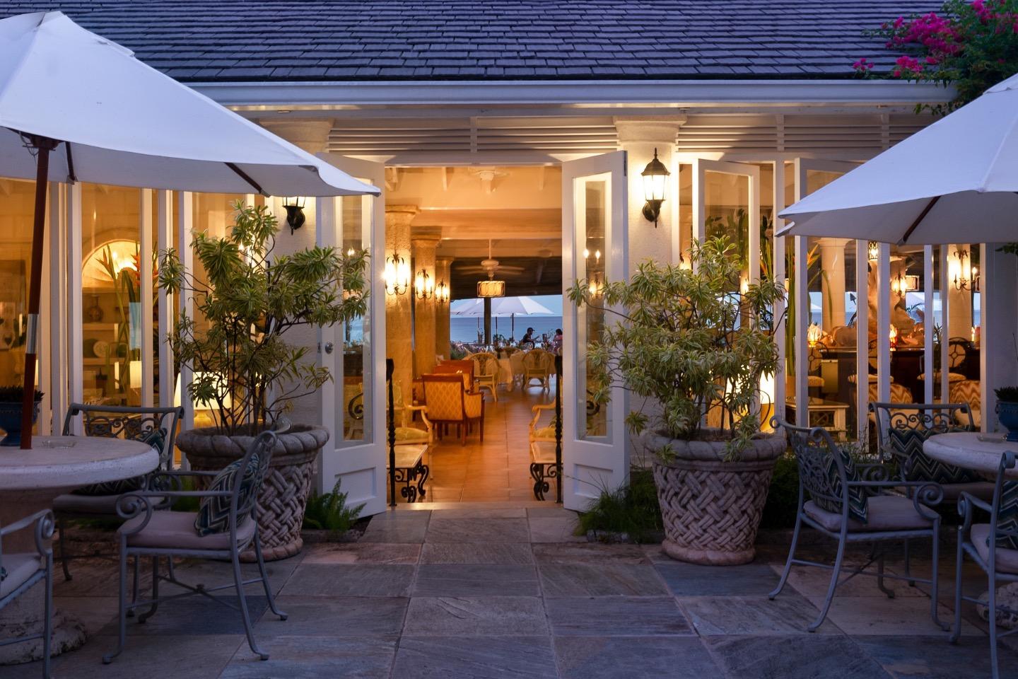 The hotel's entrance at dusk, lit doorways and lanterns under a sloped roof, with tables and umbrellas either side and a pink-flowered shrub in the corner.