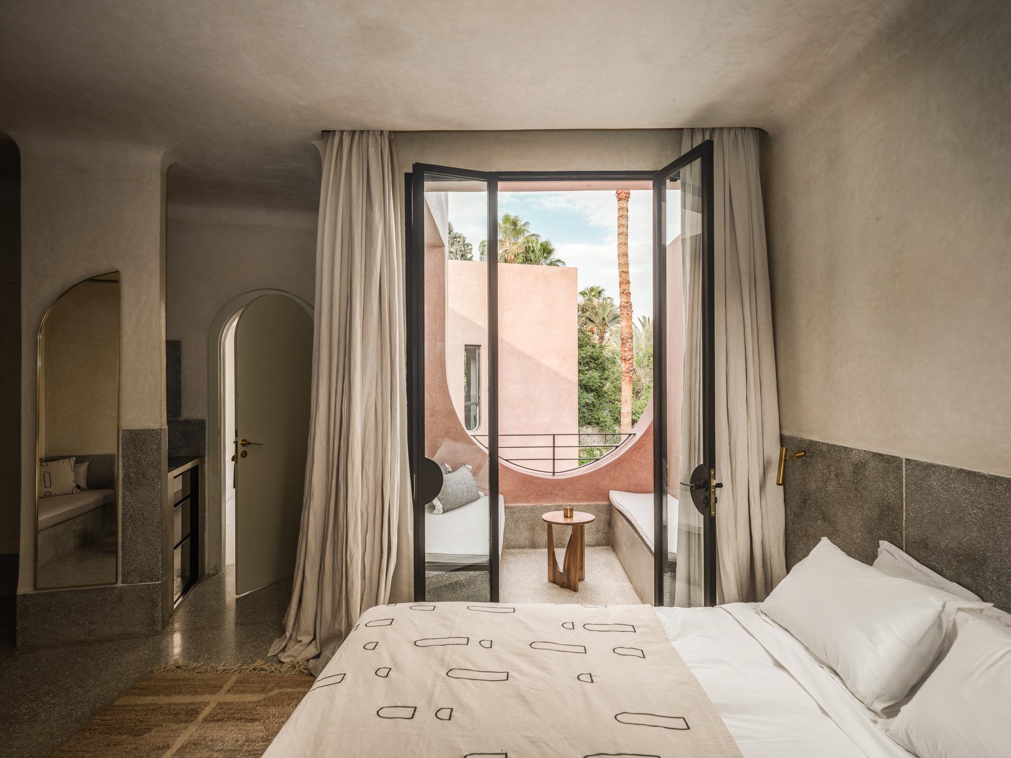 a bedroom with open glazed doors onto a balcony with built-in seating, looking out at palm trees and a pink building next door