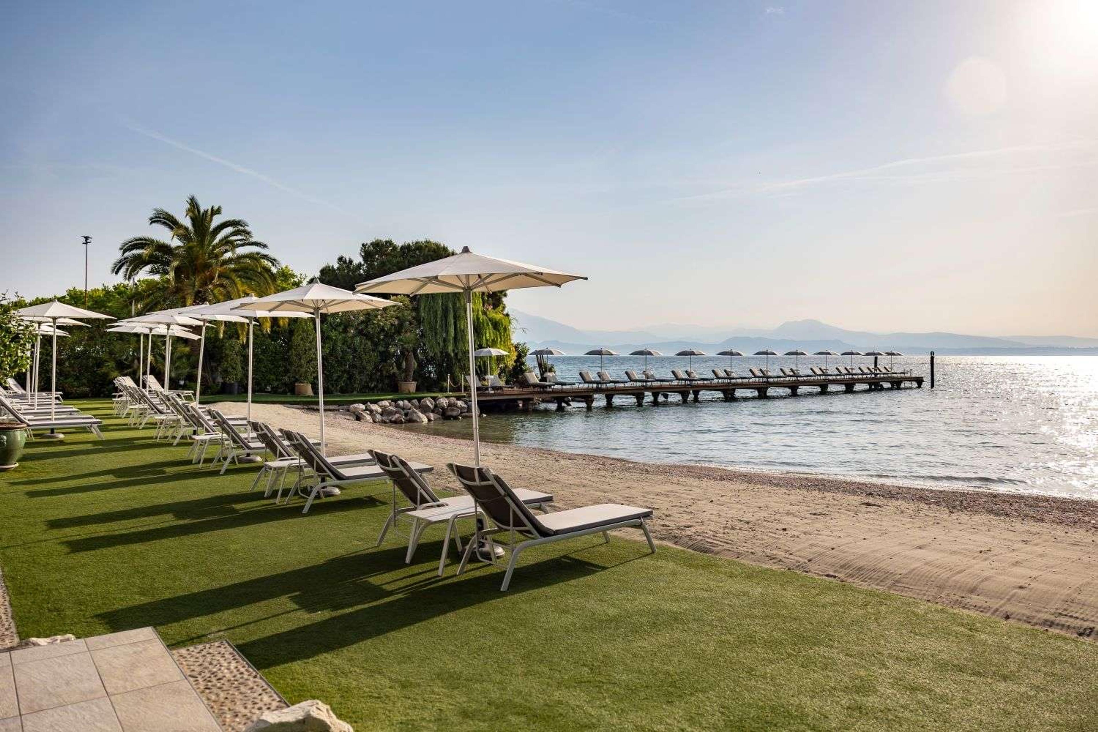 Rows of sun loungers and umbrellas on a lawn beside a pebble beach, with a long wooden dock reaching into the lake.