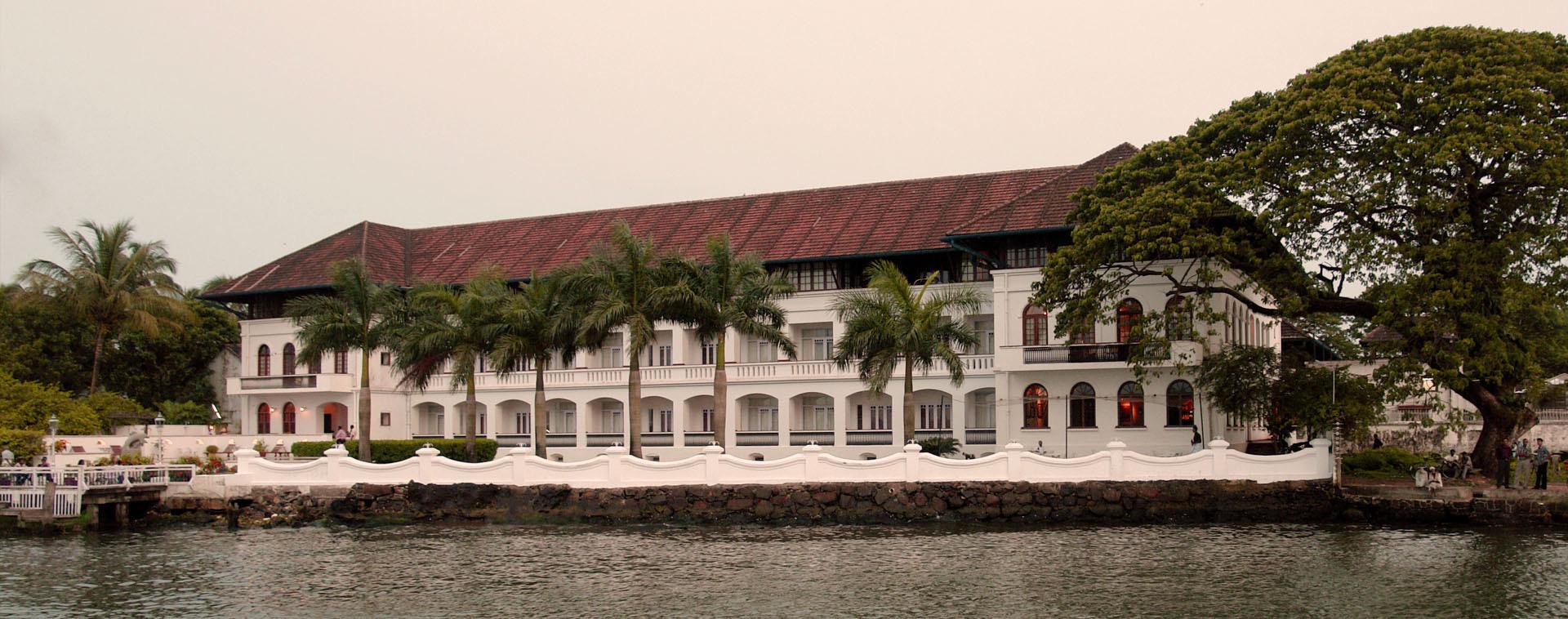 A three-storey white hotel with a red tiled roof and palm trees along its waterside terrace, seen from across the water under an overcast sky.