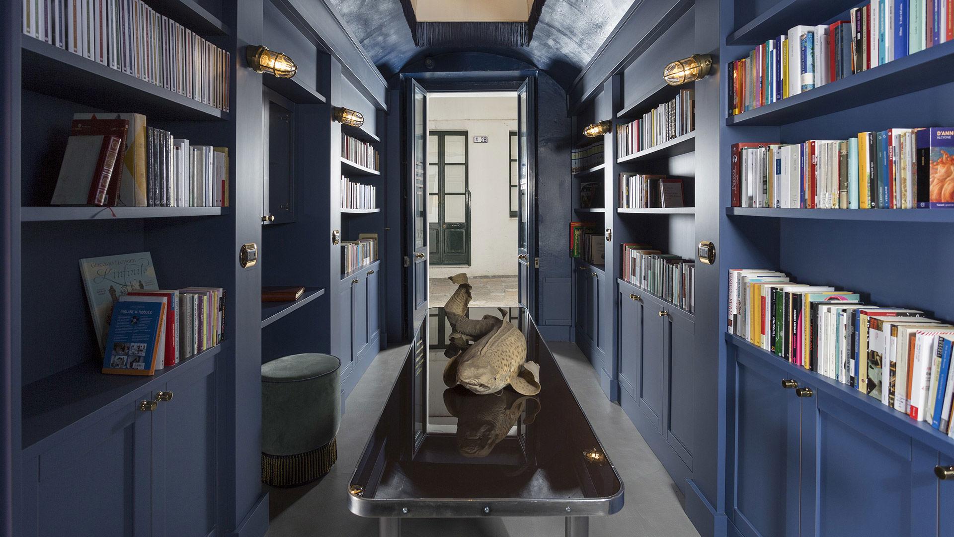 A narrow corridor lined with blue bookshelves full of books either side, a mirrored metal table holding a carved fish sculpture, leading to an open door onto the street.