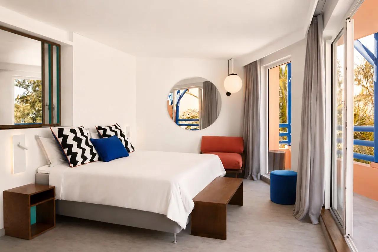 A hotel room with an orange armchair, a round mirror and a blue stool, with doors open to a balcony.