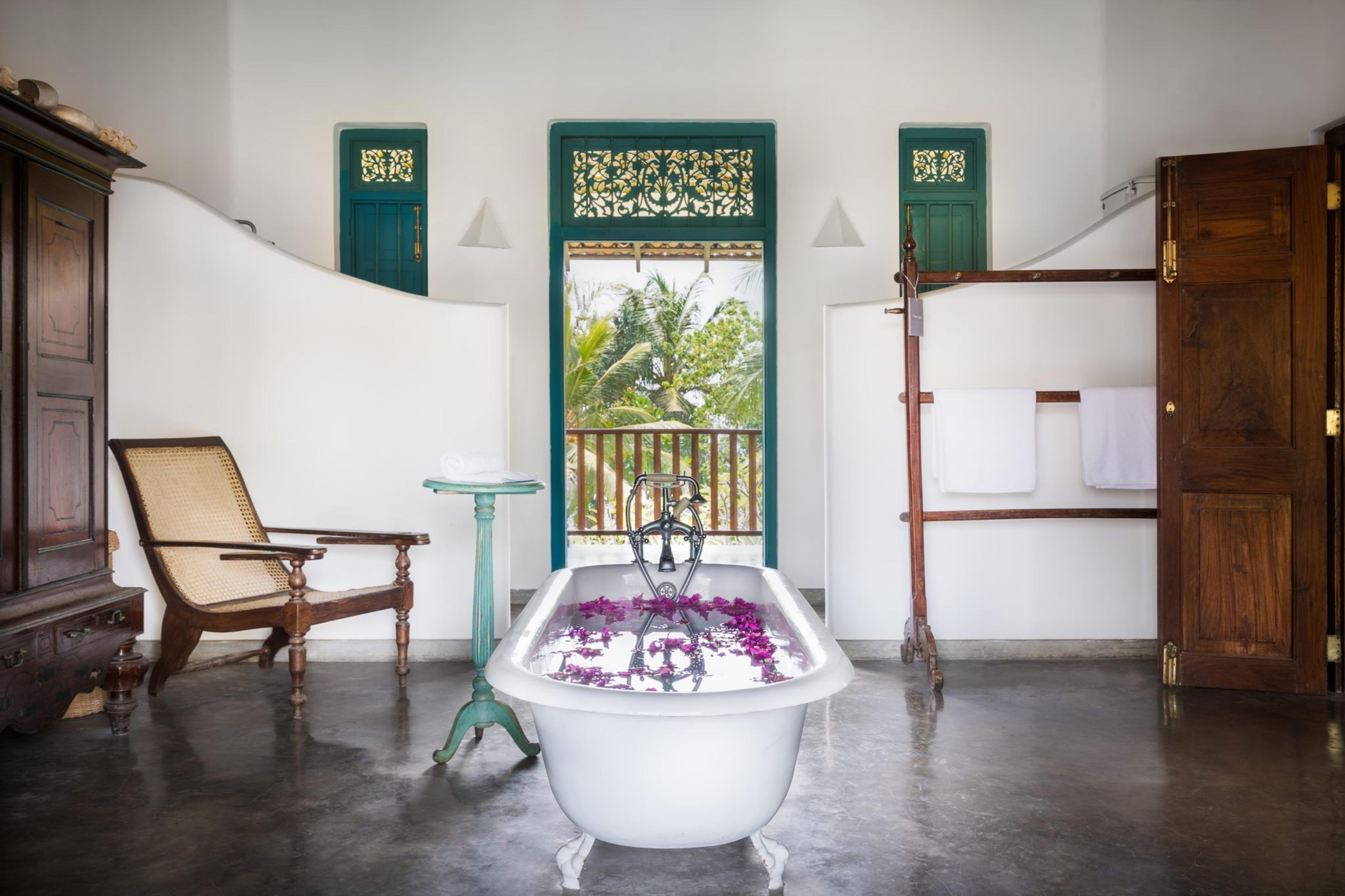 A white clawfoot bathtub scattered with pink petals stands in an open bathroom with a cane chair, a turquoise door and a wooden towel rail, palm trees visible through the doorway.