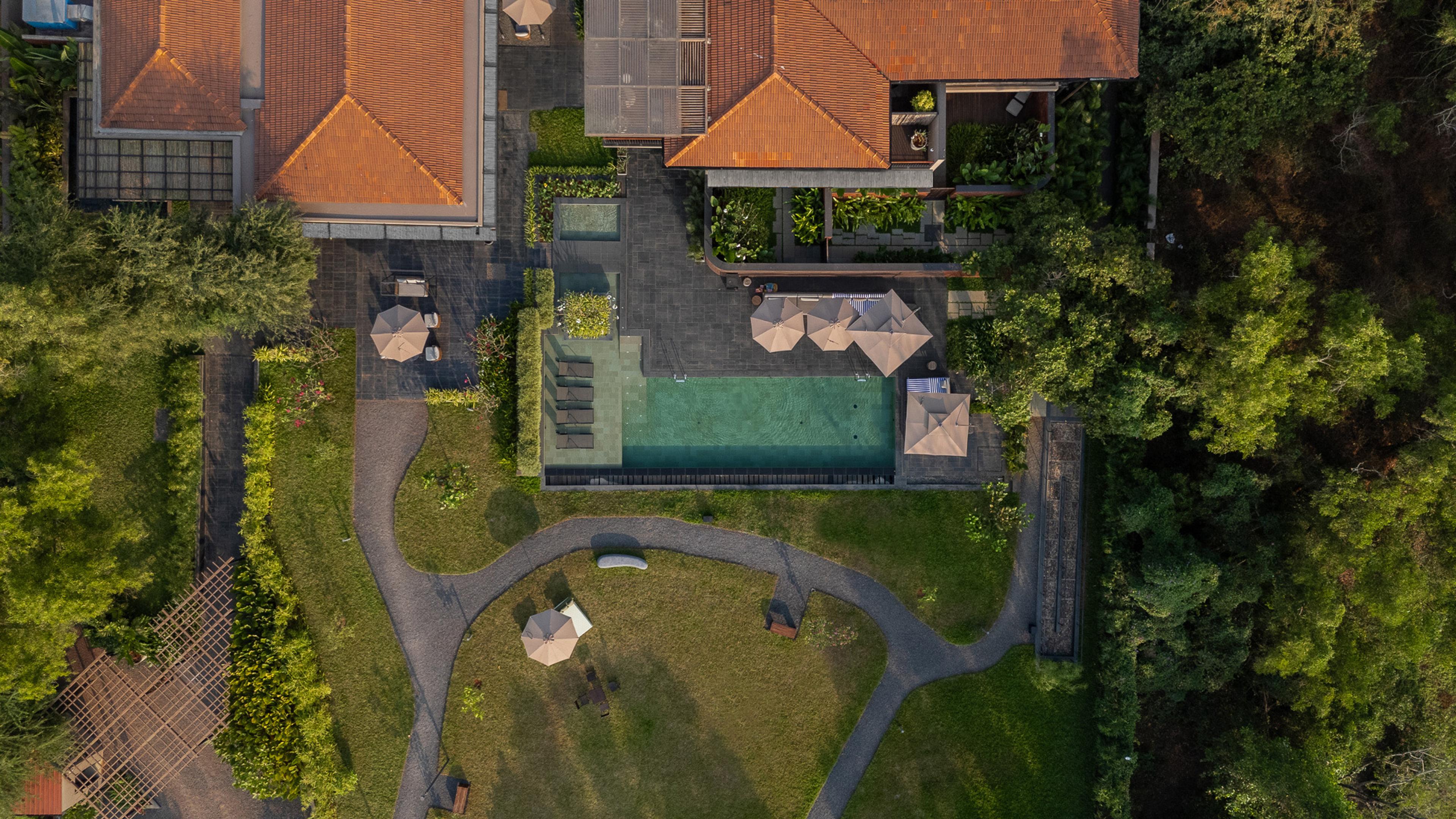 A drone view straight down on a tiled-roof building, a swimming pool, sun loungers and umbrellas, surrounded by lawn, pathways and trees.