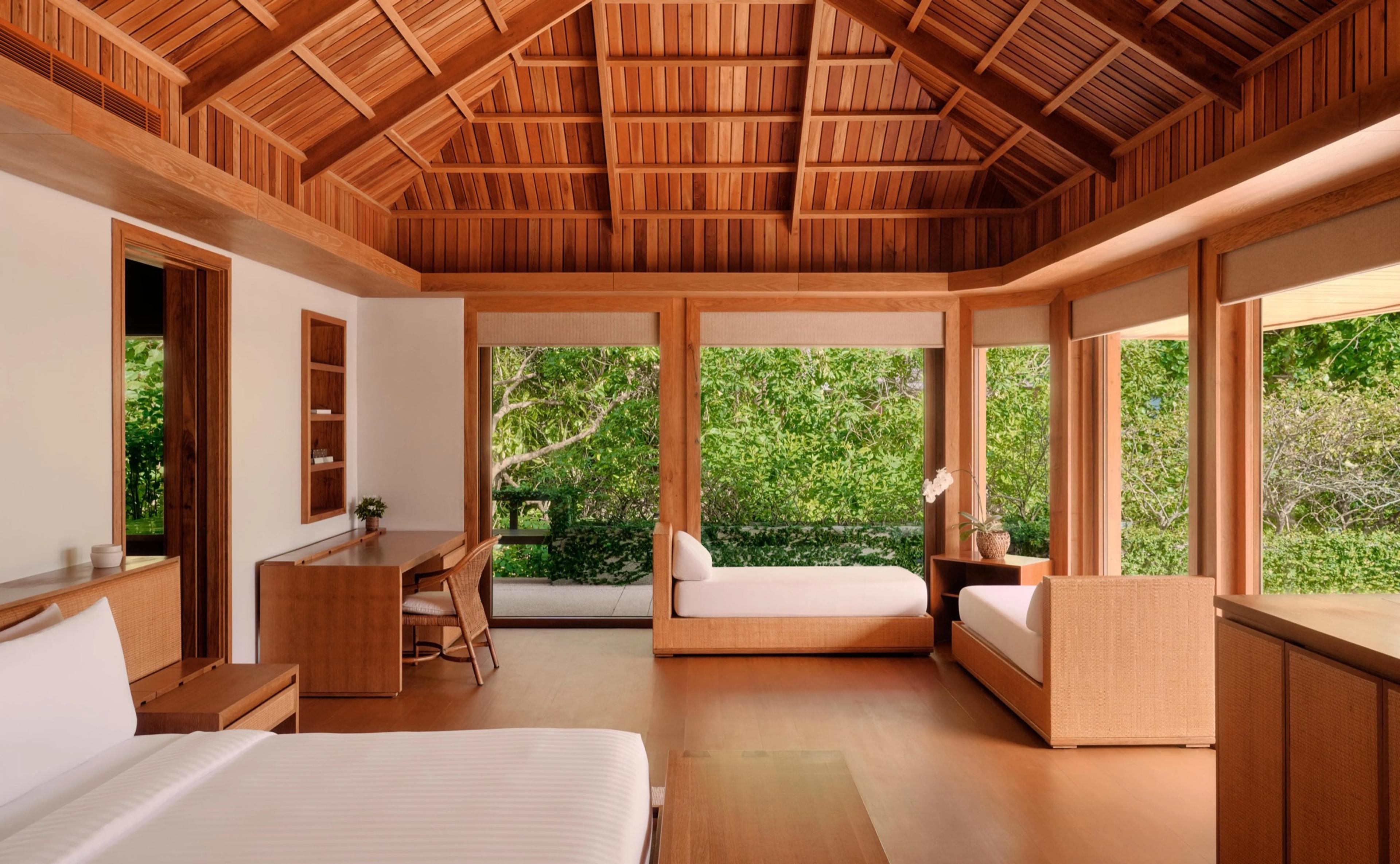 A bedroom with a peaked wooden ceiling, a bed in the foreground, and a daybed and sofa by floor to ceiling windows looking onto trees.