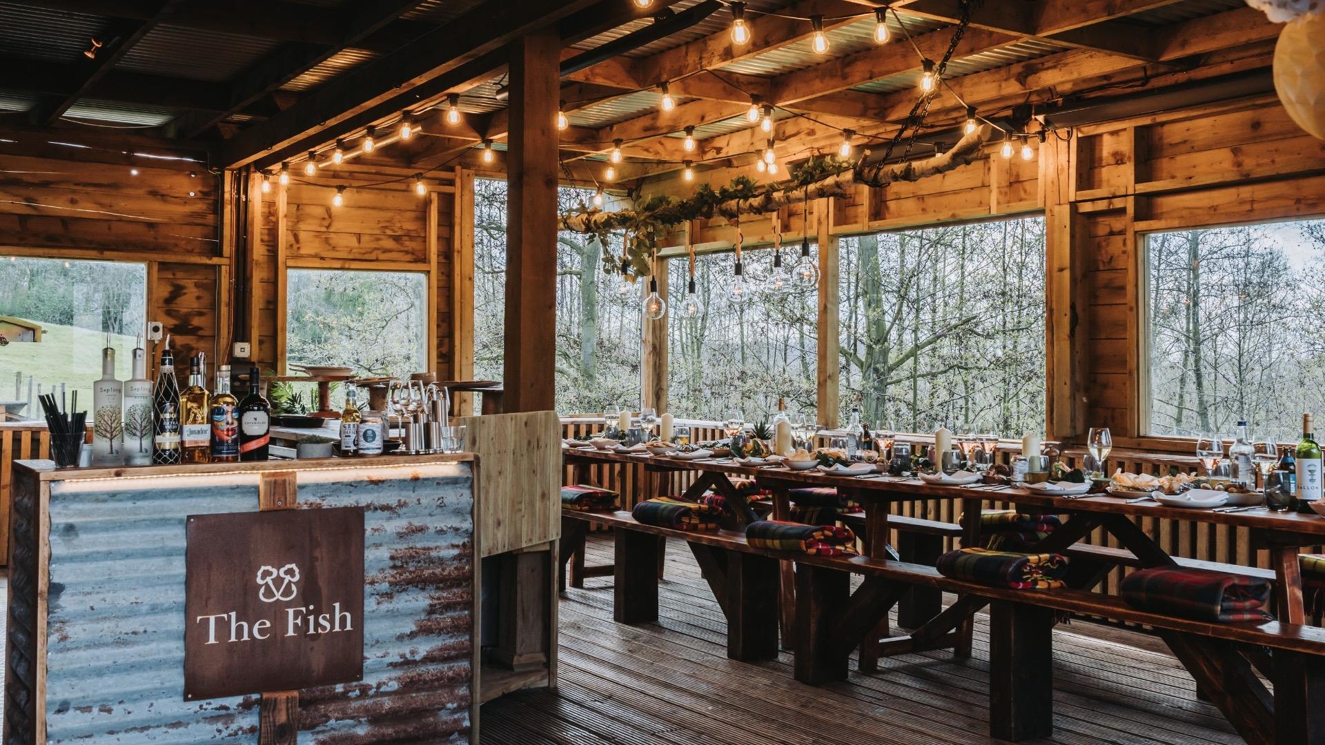 An open-sided wooden barn strung with lights, laid with long benches and tables set for a group, with a sign reading The Fish over the bar and forest visible beyond.
