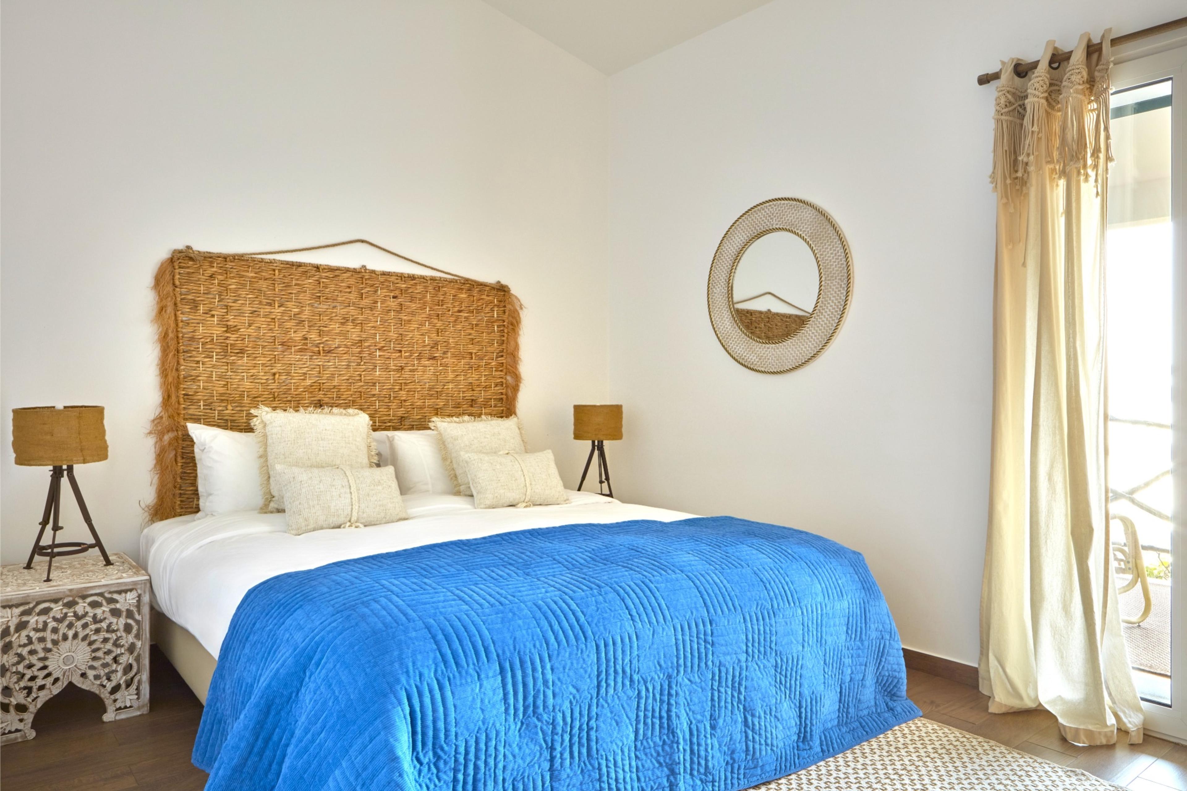 A bed with a woven rattan headboard, white pillows and a bright blue quilt, a round rattan mirror on the wall and cream curtains beside a glass door.