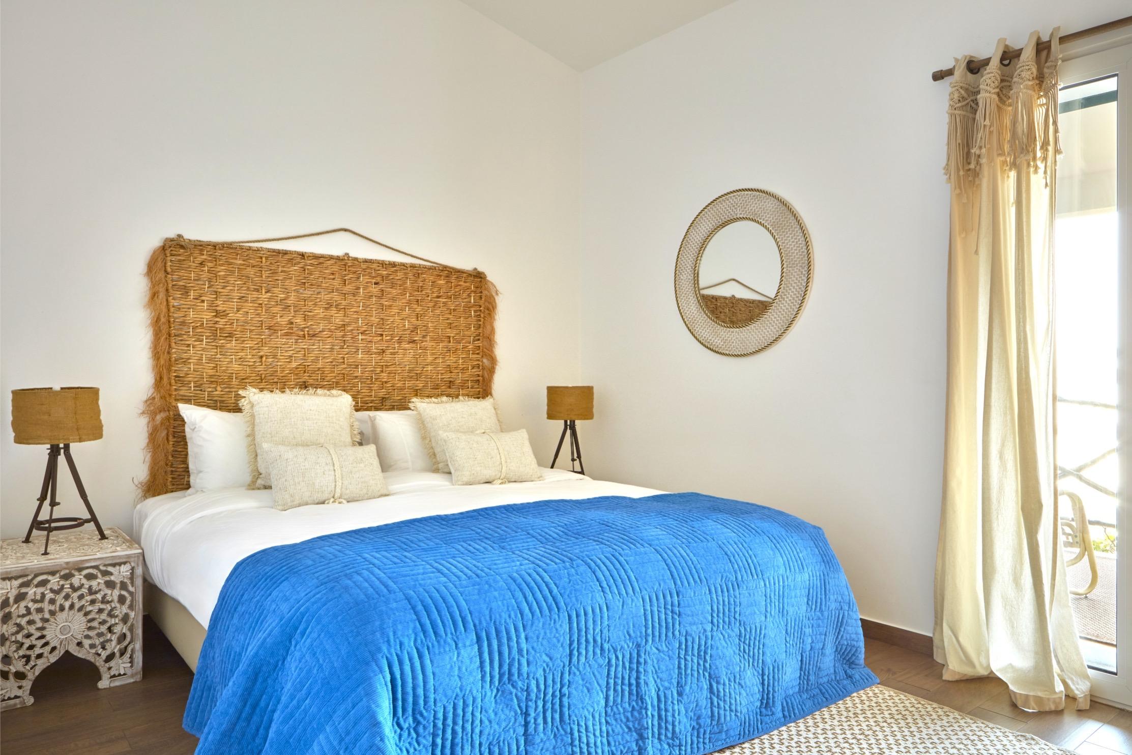 A bed with a woven rattan headboard, white pillows and a bright blue quilt, a round rattan mirror on the wall and cream curtains beside a glass door.
