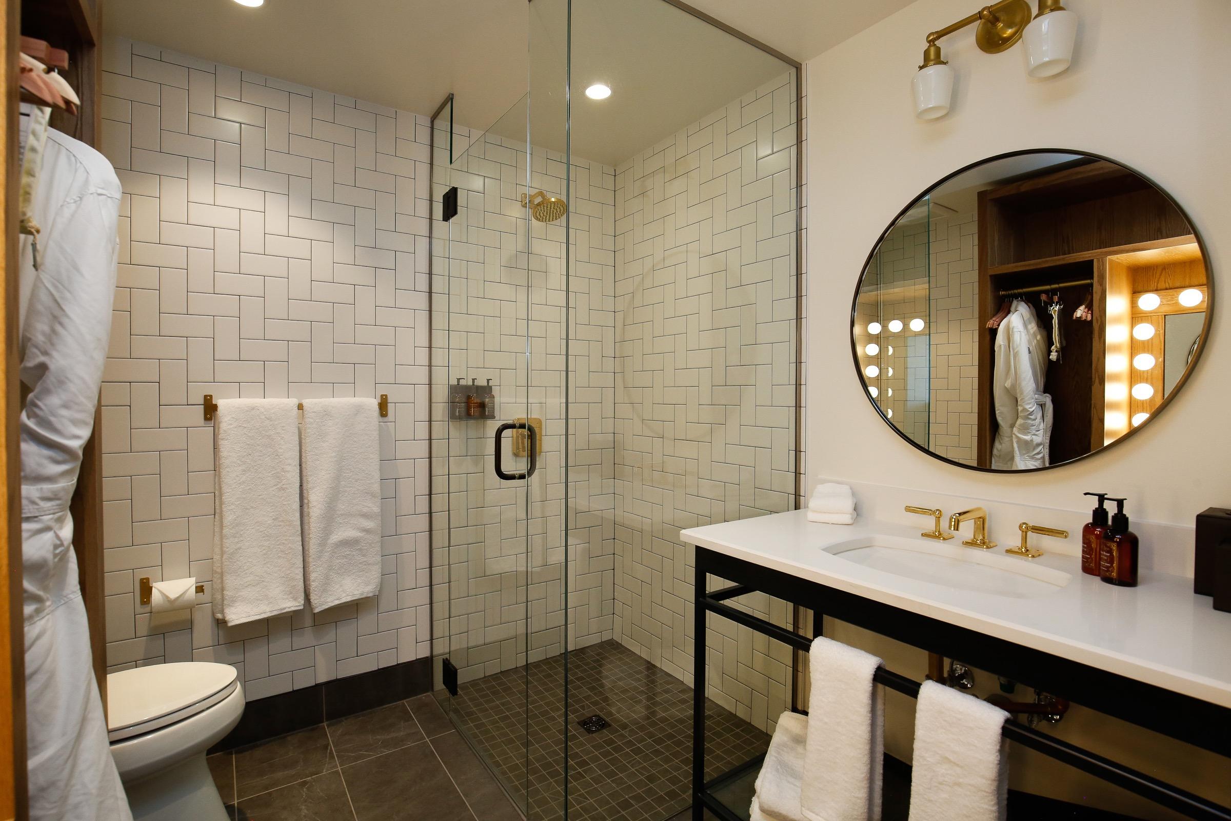 A bathroom with a glass walk in shower, herringbone tile, a white robe hanging beside it, and a round mirror over a double vanity.