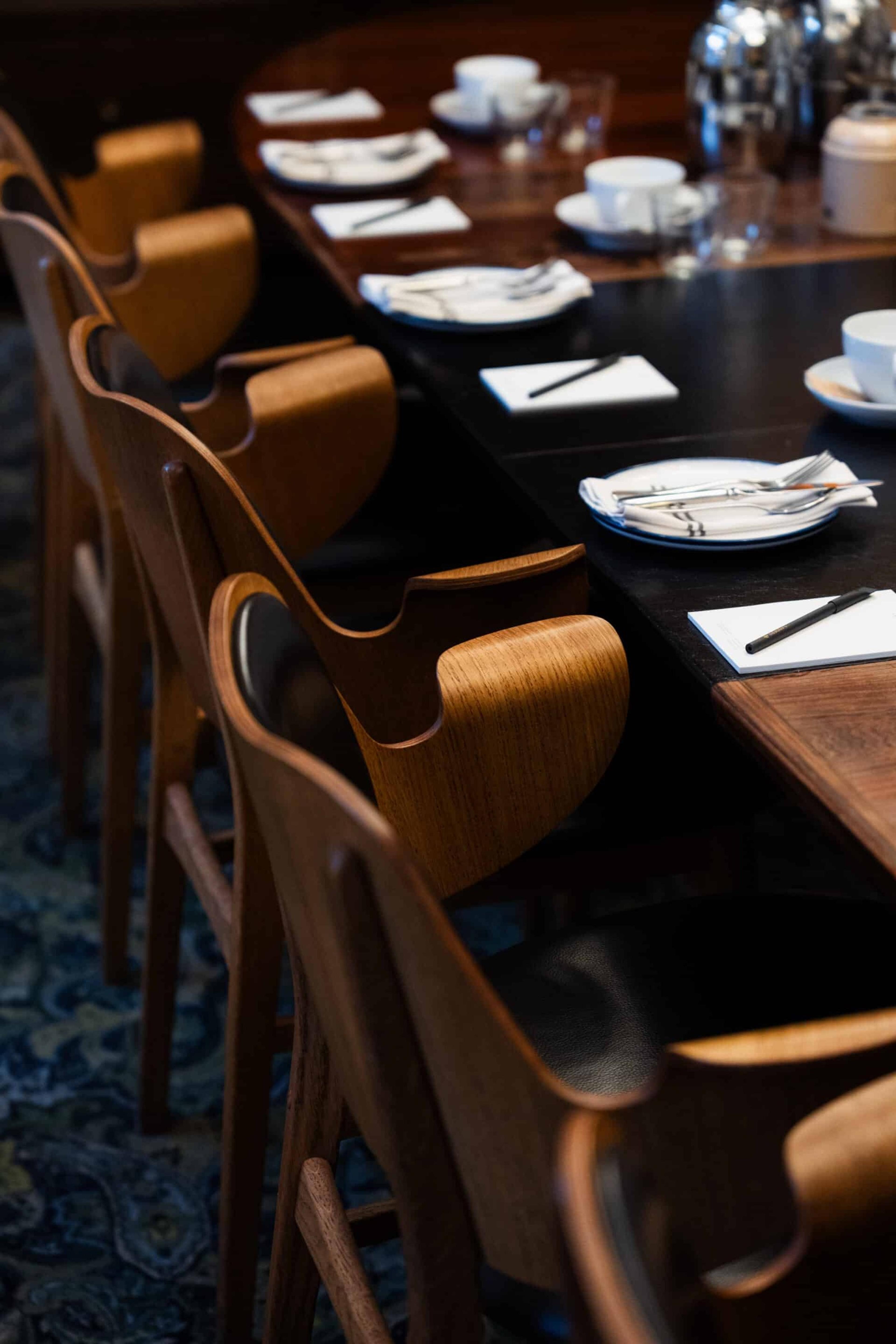 A long dark wood table laid with white napkins, cutlery, notepads and pens, bordered by a row of curved wooden chairs on a patterned carpet.