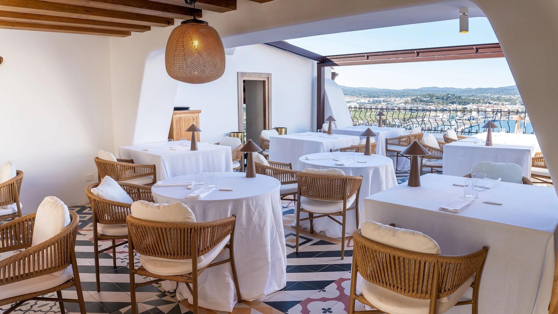 A covered restaurant terrace with tables laid in white cloths and woven armchairs, open on one side to a bay and a town on the hills.