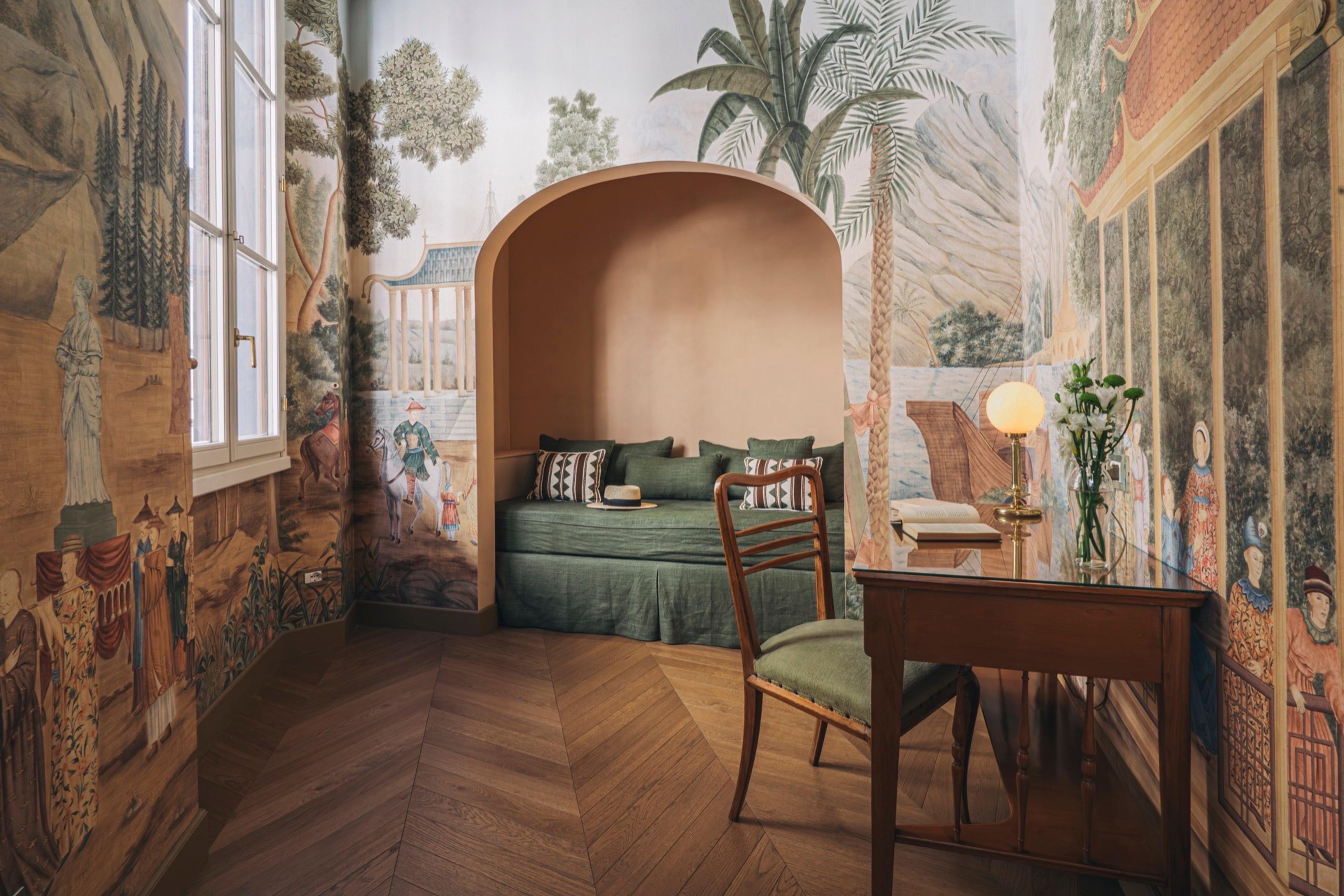 A daybed with green cushions sits in a plastered alcove surrounded by walls hand-painted with a classical landscape mural, a writing table and chair in front.