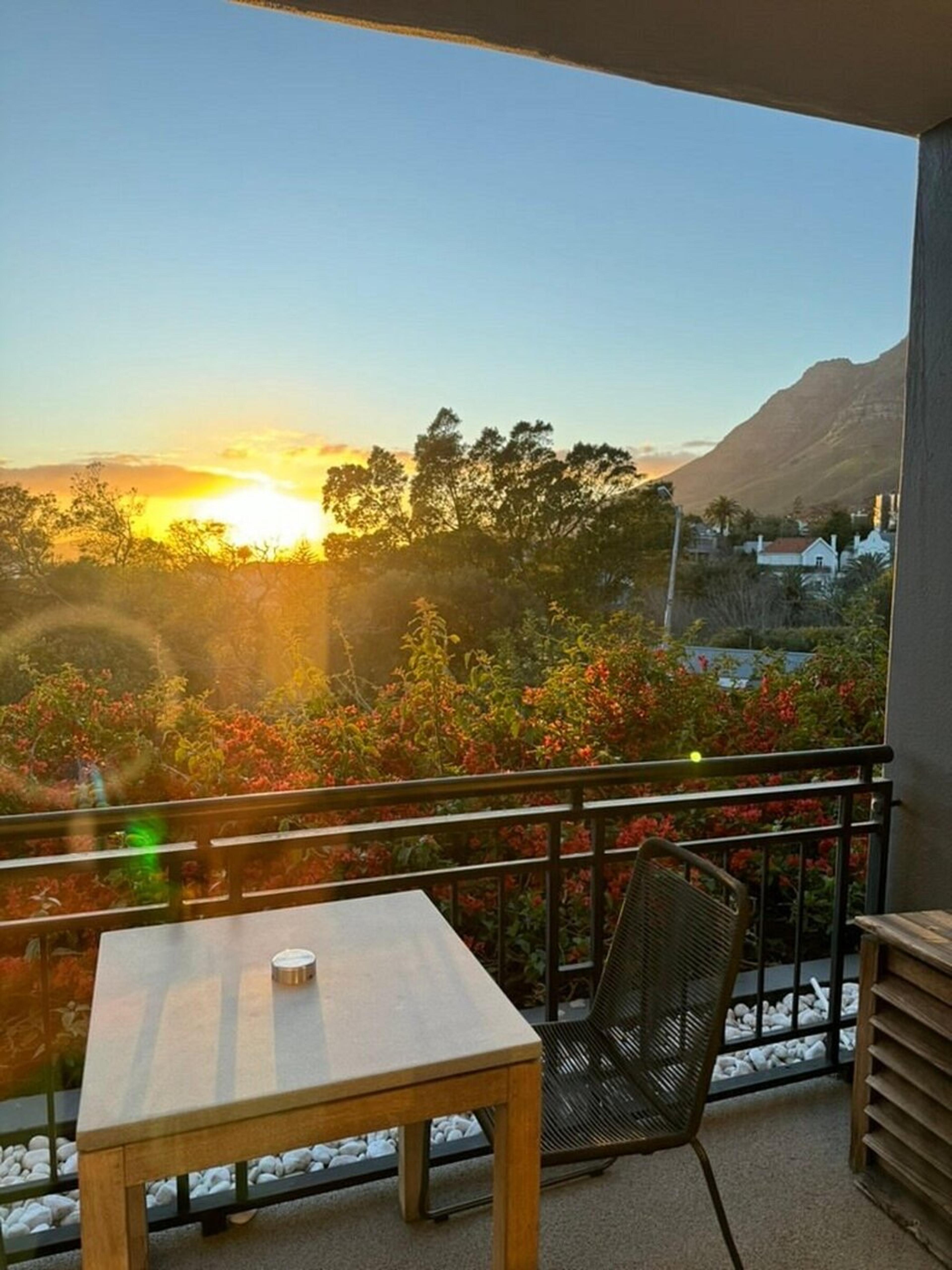 A small balcony at sunset with a square table and a woven chair, a metal railing with red-flowered shrubs below, and a mountain ridge in the distance.