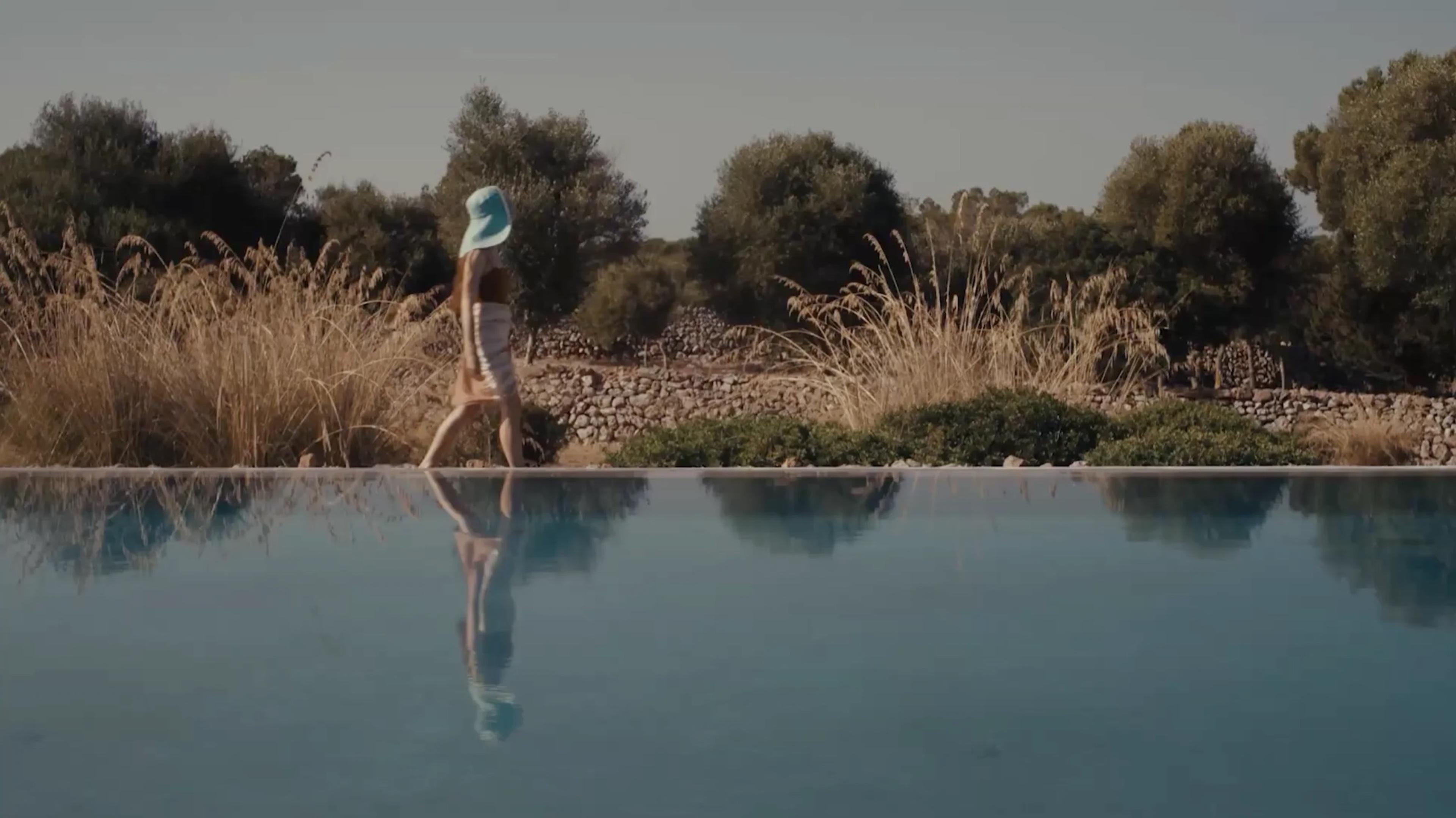 A woman in a wide-brimmed hat walks beside a still pool, her reflection in the water, with dry grasses and a stone wall behind.