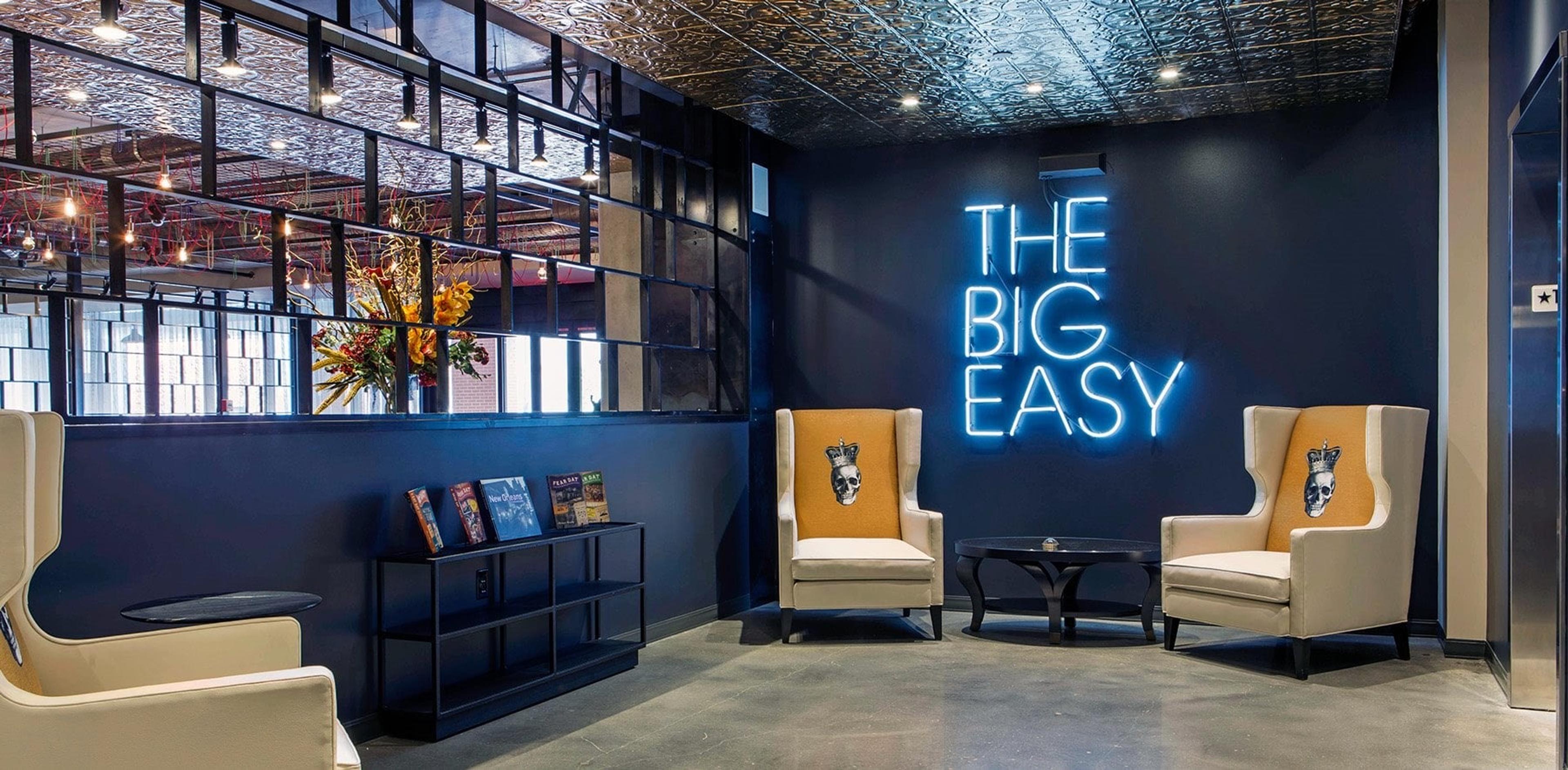 A lounge with two wingback chairs, a neon sign reading THE BIG EASY on a dark blue wall, and a pressed-tin ceiling.