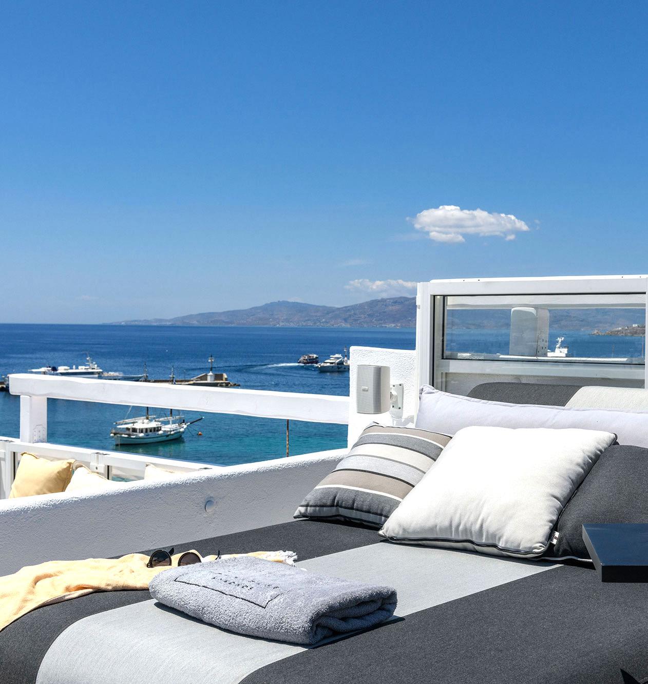 A sun lounger with pillows, a folded towel and sunglasses, overlooking a harbour with boats on the sea.