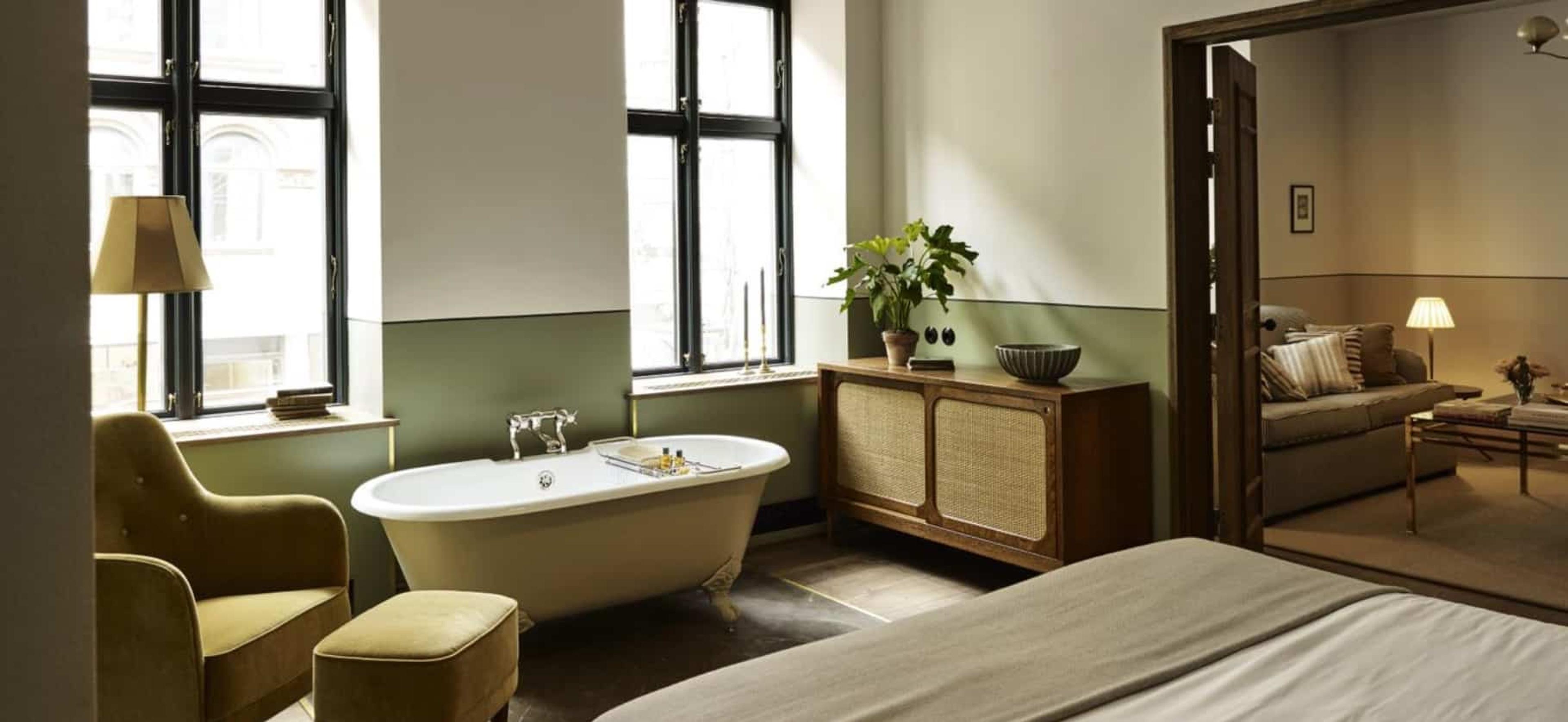 A freestanding tub beside tall windows in a green and white bedroom, with a velvet armchair and ottoman in the foreground and a sitting room visible through an open door.