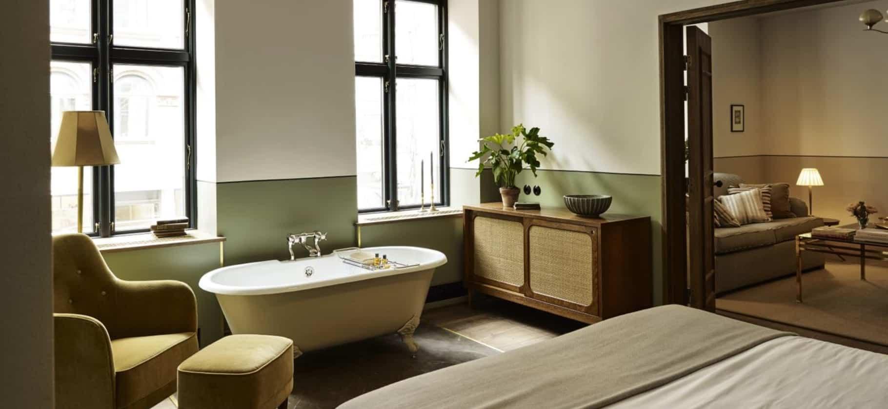 A freestanding tub beside tall windows in a green and white bedroom, with a velvet armchair and ottoman in the foreground and a sitting room visible through an open door.