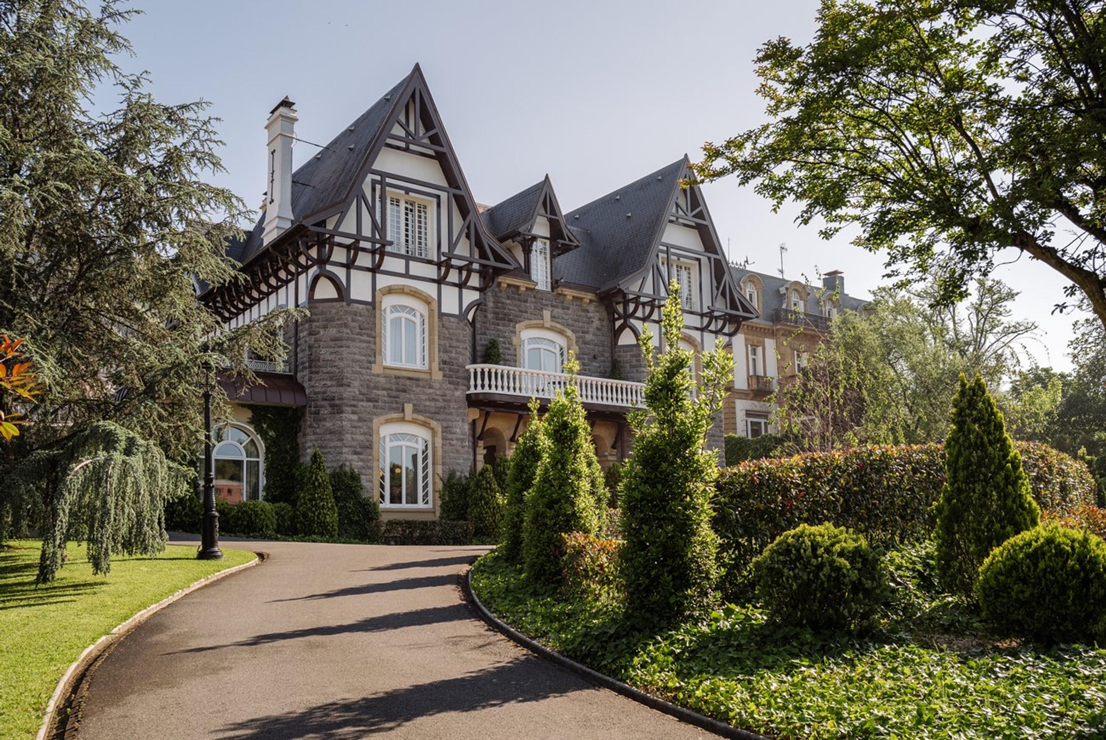 A stone and timber villa with three gables seen from a curving driveway lined with trees and clipped shrubs under a blue sky.