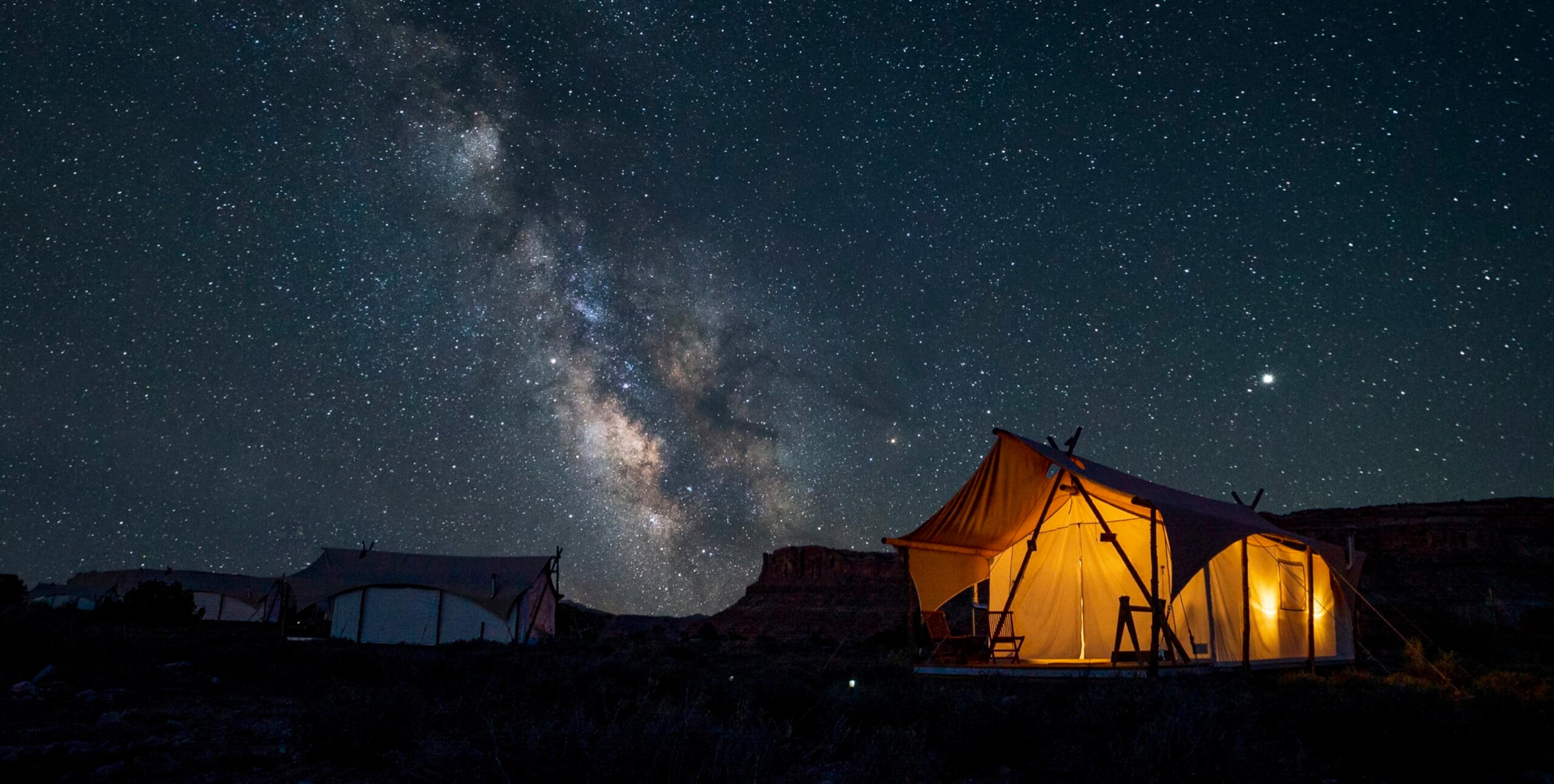 A night sky with the Milky Way over a row of canvas tents on dry ground, one tent lit from inside, with a flat-topped red rock butte behind them.