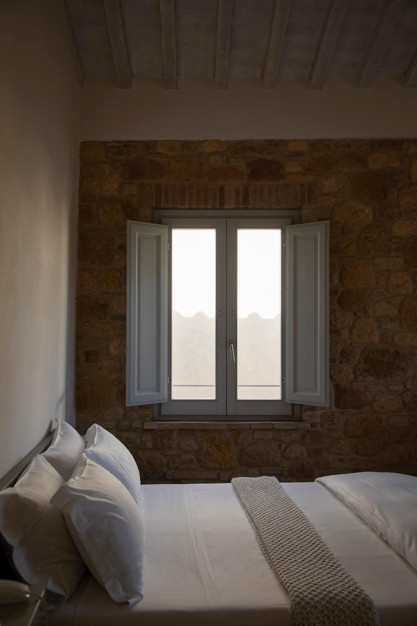 A bed with white pillows against a stone wall, below an open shuttered window looking out to the sky.