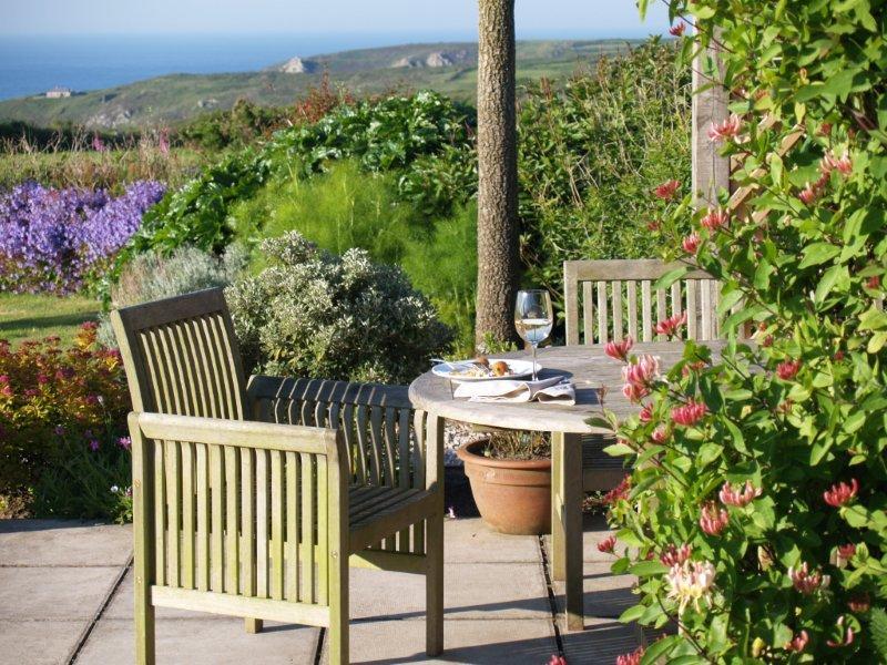 A table and two wooden chairs in a flowering garden, with the moor and the sea beyond.